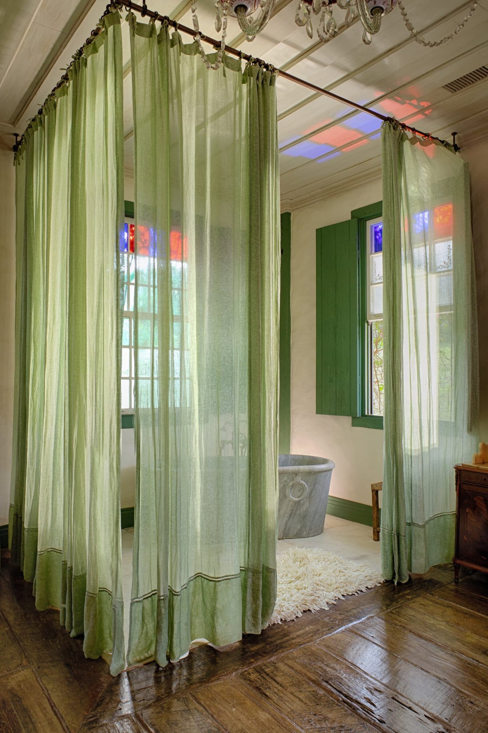 Leinenstoff, Buntglaslicht und ein Steinbad. Mehr braucht es wirklich nicht. Photo: Henrique Mangeon Durchscheinende salbeigrüne Himmelbettvorhänge in einem Zimmer der Ibiti Project Lodge, mit freistehendem Steinbadezuber, grünen Fensterläden mit buntem Glaslicht und poliertem Hartholzboden