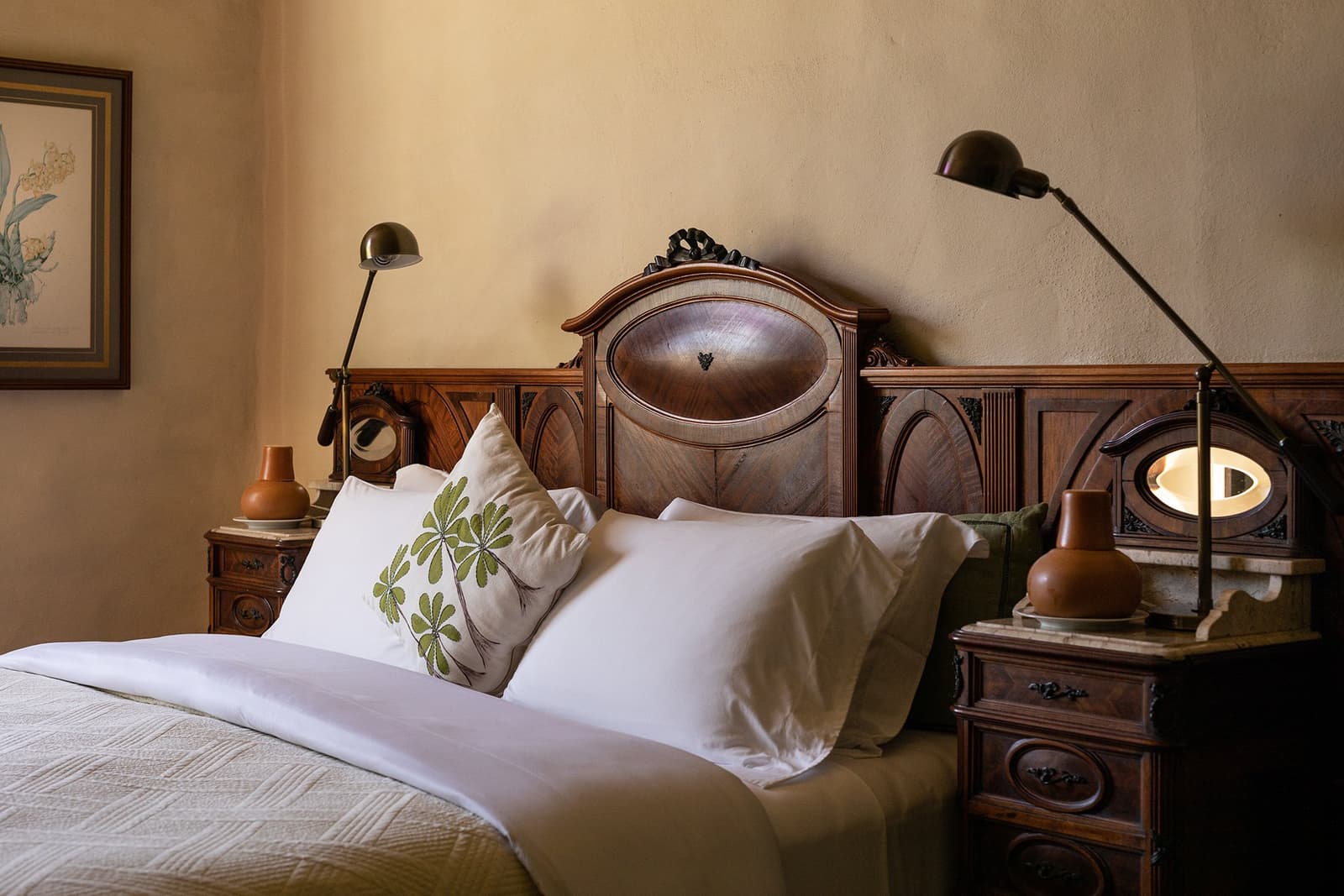 Du bois ancien, du linge frais, et un coussin qui a lu son herbier. Photo: Henrique Mangeon Un lit double avec du linge blanc immaculé et un coussin brodé à motif botanique devant une tête de lit en noyer sculpté à l'Engenho Lodge de l'Ibiti Project, encadrée de chevets anciens assortis et de lampes de lecture en laiton