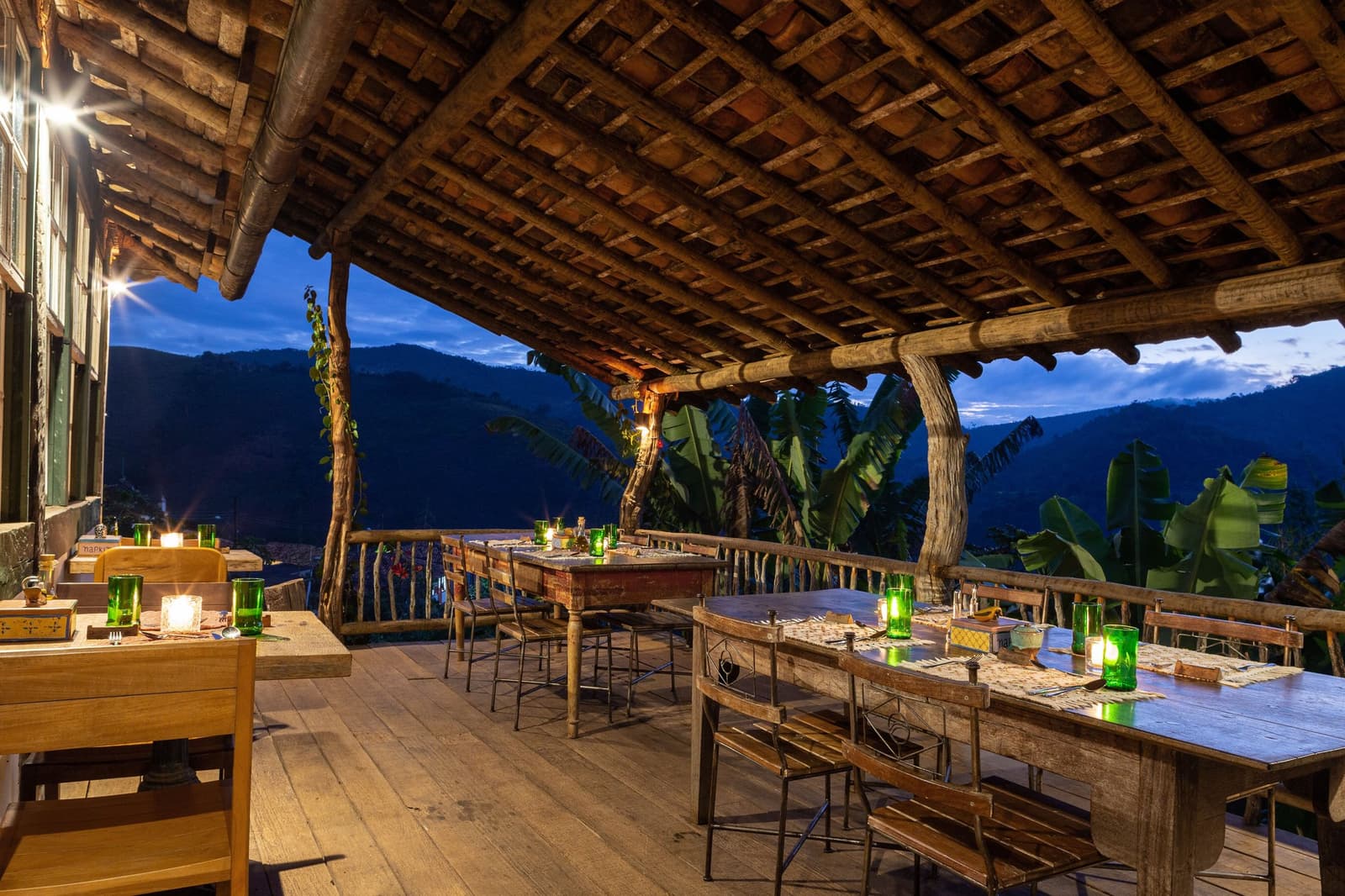 Das Abendessen steht, das Tal hat seine Lichter gedimmt, und die Berge übernehmen den Rest. Kerzenbeleuchtete Außenterrasse der Engenho Lodge des Ibiti Project, mit rustikalen Holztischen zum Abendessen unter einem Balkenholzdach und einem Bergtal in der Abenddämmerung dahinter