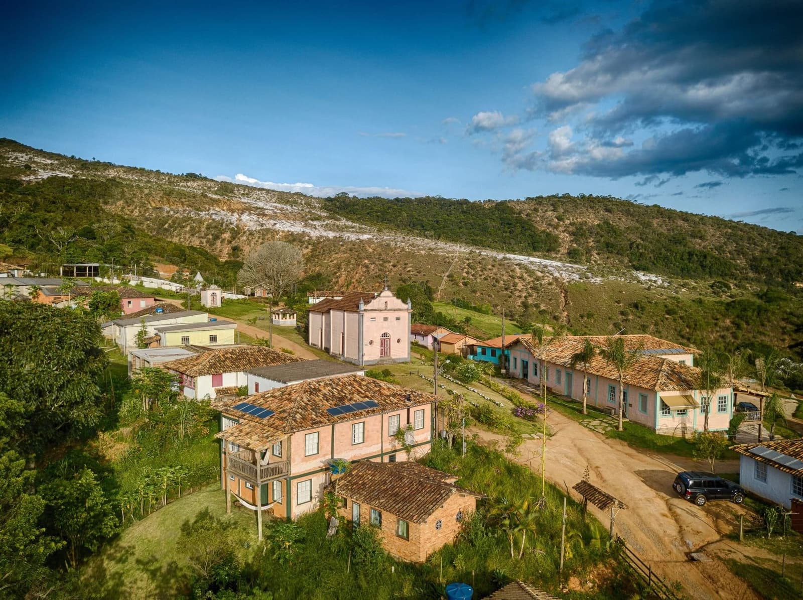 Un village où le temps a ralenti, sans jamais s'arrêter. Vue aérienne d'un petit village historique au Minas Gerais, avec des maisons coloniales aux teintes pastel, des toits en tuiles de terre cuite, une chapelle rose, un chemin de terre et des collines boisées avec des affleurements rocheux blancs en arrière-plan, au sein de la réserve de l'Ibiti Project.