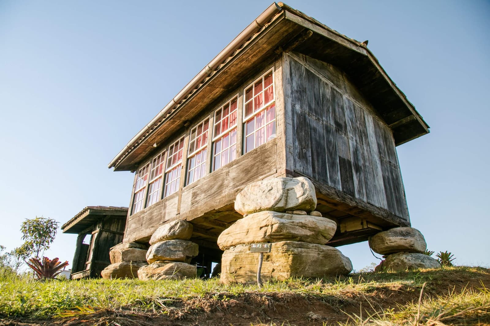 Construit pour durer, bien avant qu'on parle de patrimoine. Un bâtiment en bois patiné par le temps, surélevé sur des fondations de pierres empilées dans la réserve du Projet Ibiti au Minas Gerais, photographié en contre-plongée sous un ciel bleu limpide, avec des rideaux rouges visibles à travers les fenêtres à petits carreaux