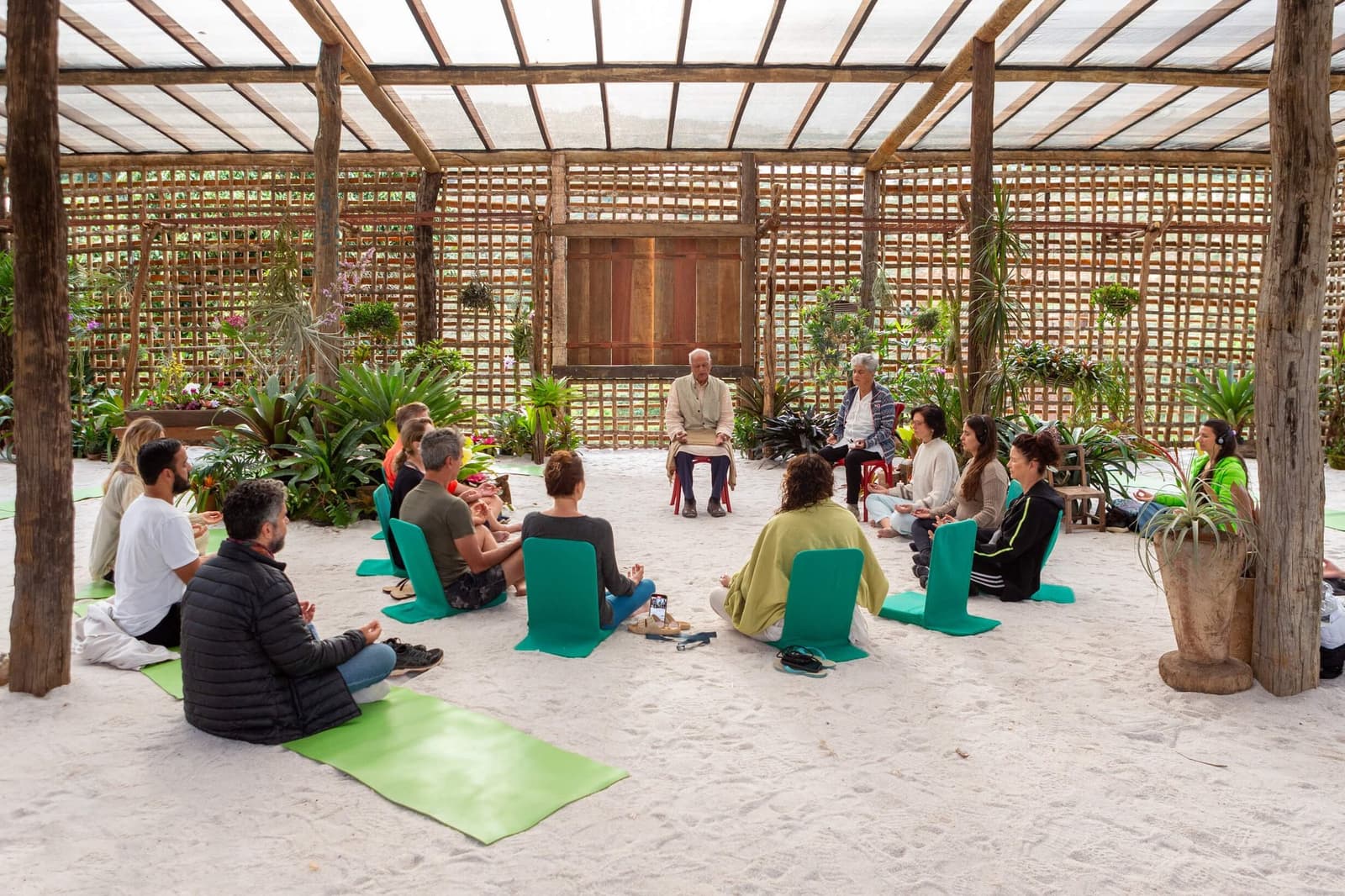 Le sable, le silence, et des orchidées qui font le décor. Un groupe de voyageurs assis en cercle sur du sable blanc à l'intérieur d'un pavillon en bambou ouvert entouré de plantes tropicales, lors d'une séance de méditation guidée à l'Ibiti Project au Minas Gerais.