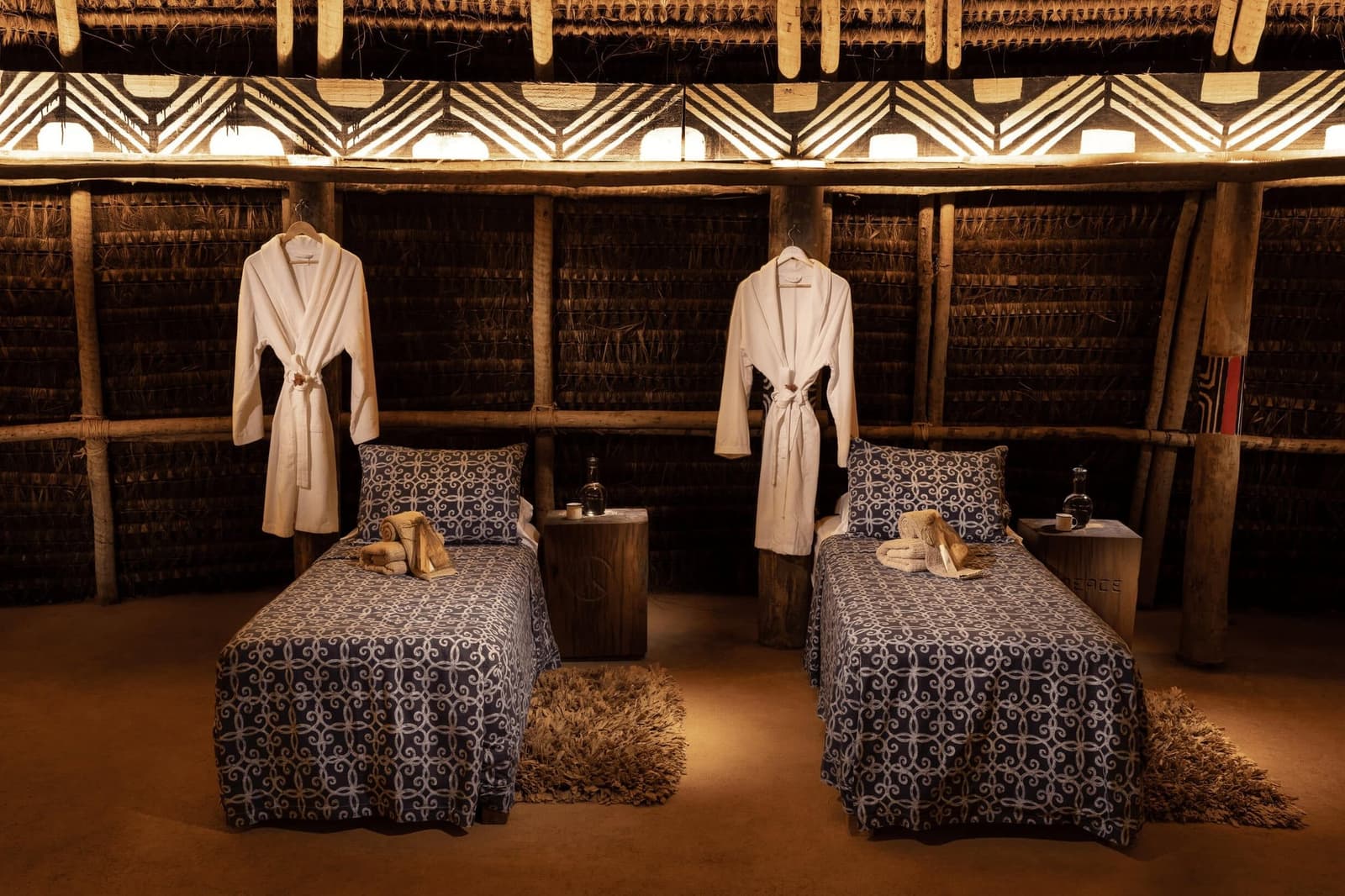 Peignoir prêt, la forêt s'occupe du reste. Deux lits de massage avec du linge à motifs bleus et des peignoirs blancs accrochés à des poteaux en bois dans une Oca au toit de chaume du Ibiti Project, éclairée d'une lumière ambrée