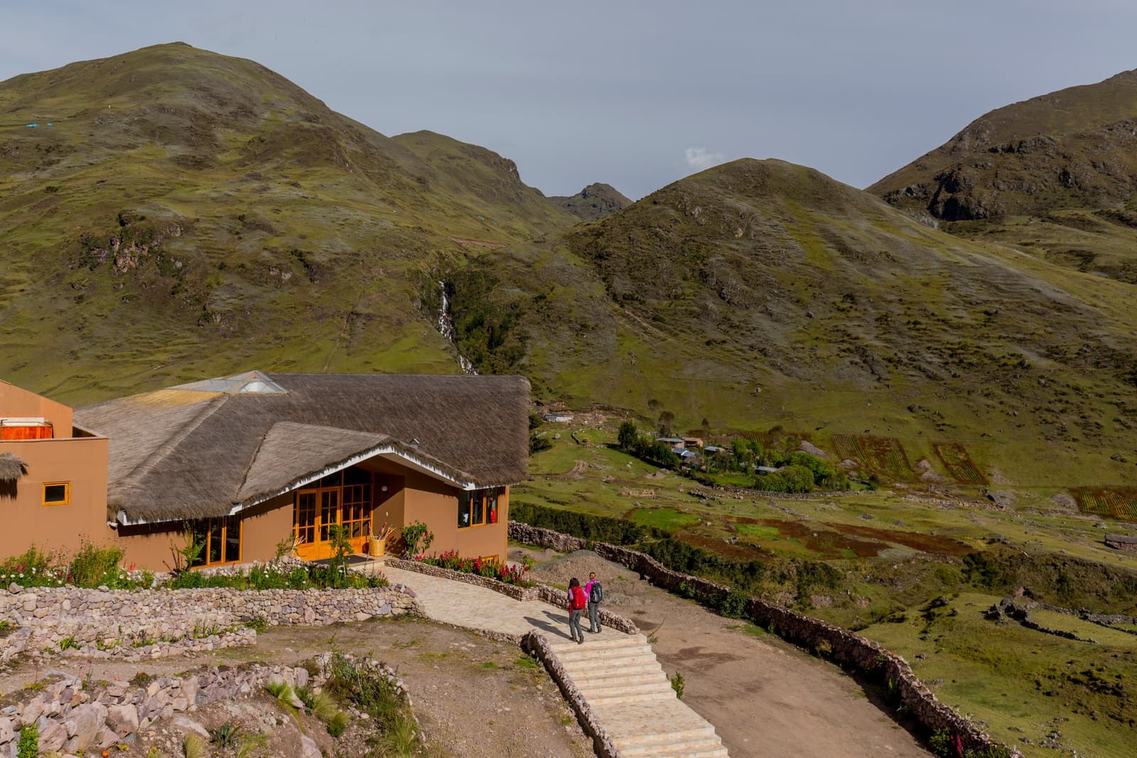 Your room has a hot tub. The waterfall outside comes free. A thatched-roof mountain lodge with warm ochre walls and yellow-framed windows sits on a hillside on this luxury Machu Picchu tour, as two guests walk a stone-lined path toward the entrance, with green Andean peaks and a distant waterfall behind