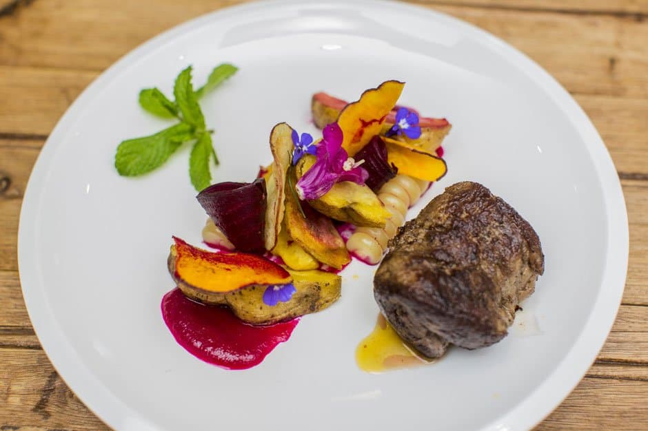 Dinner in the Andes takes its presentation seriously, and rightly so. A plated dish on a luxury Machu Picchu tour featuring a seared meat medallion, roasted root vegetable chips, beet purée, edible blue and pink flowers, and fresh mint on a white ceramic plate set on a wooden table