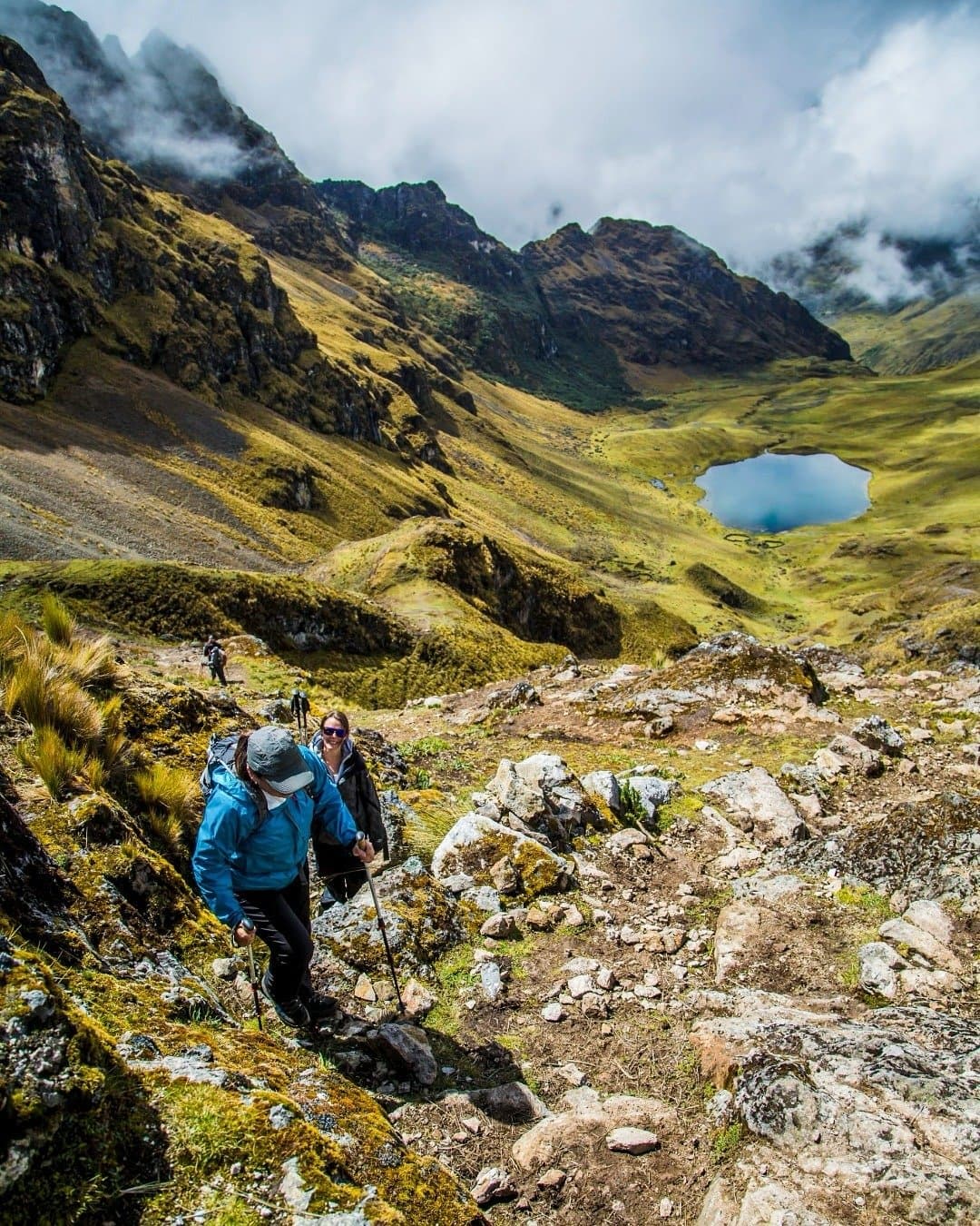 That lake in the valley below? You earned it, step by step. Two hikers with trekking poles climbing a rocky trail on this luxury Machu Picchu tour, with a vivid blue alpine lake, green puna grasslands, and cloud-draped Andean peaks stretching into the valley behind them
