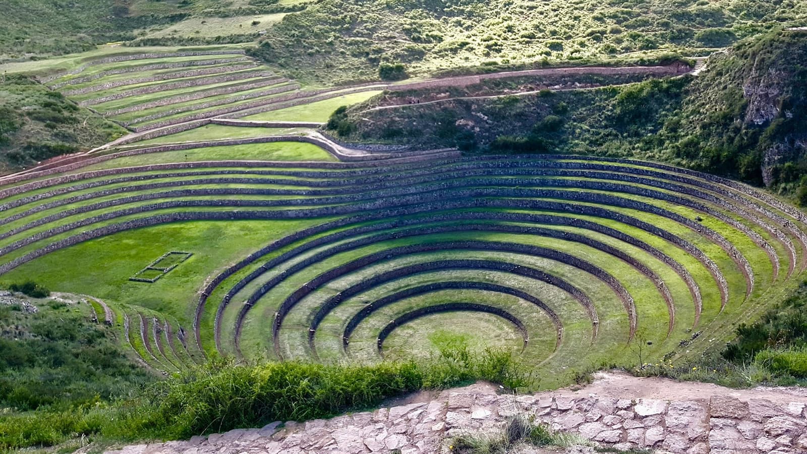 Concentric rings of green, carved into the earth by hand. The Inca had a flair for the dramatic. Aerial view of the circular Inca farming terraces of Moray, a highlight of any luxury Machu Picchu tour, with concentric stone-walled rings of vivid green grass descending into a natural depression in the Sacred Valley highlands