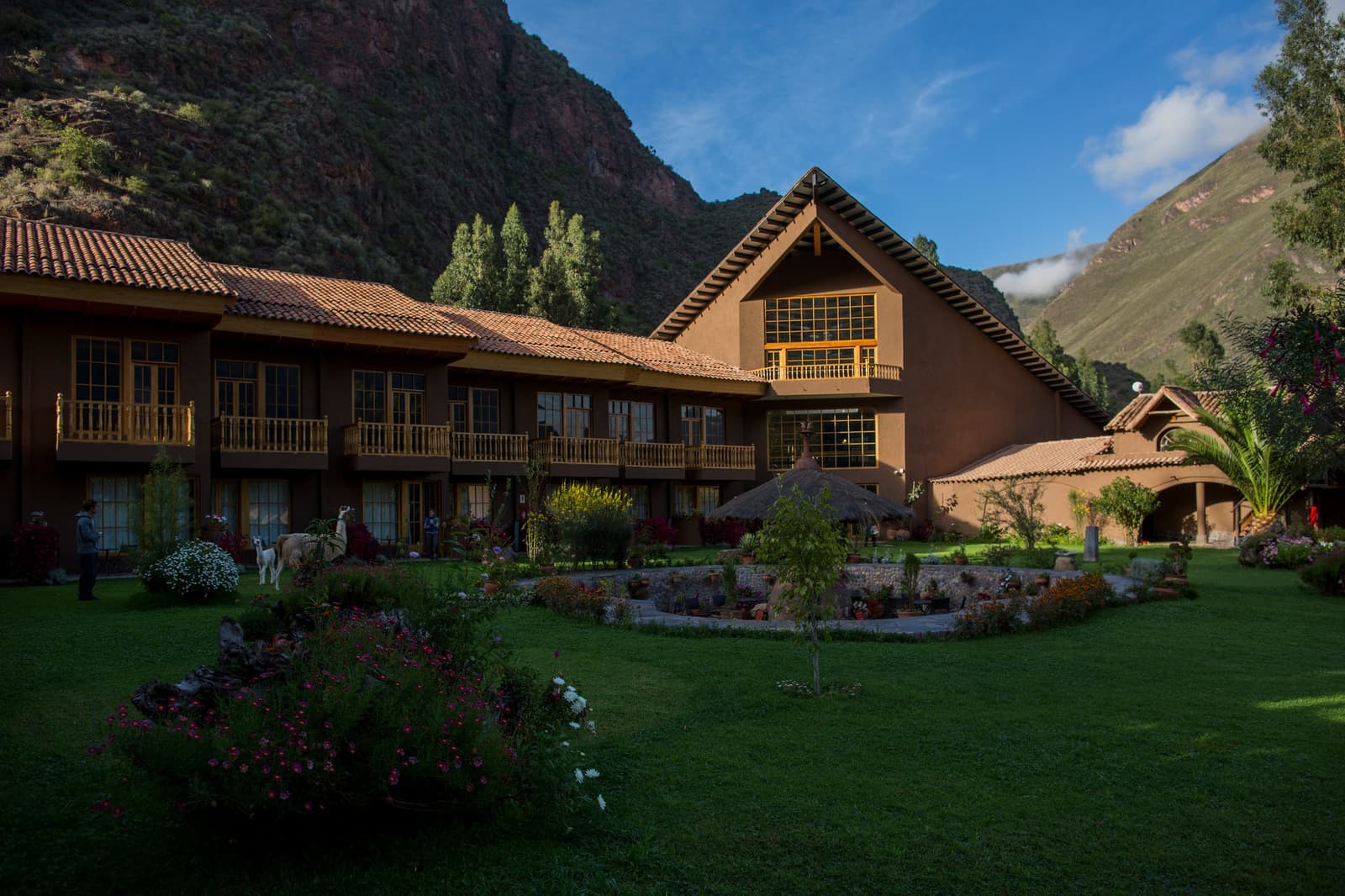 A llama in the garden keeps things real, no matter how comfortable the room. A two-storey Andean lodge with terracotta-tiled roof and wooden balconies set against steep green mountains on this luxury Machu Picchu tour, with a white llama grazing on the garden lawn and flowering beds in the foreground