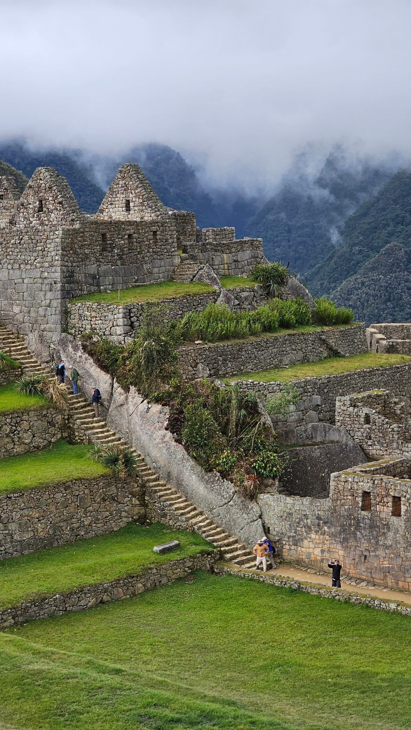 Clouds roll in uninvited, and honestly, they improve the mood. Layered Inca stone terraces and stairways at Machu Picchu on a luxury Machu Picchu tour, with low clouds drifting over the forested mountains behind the ancient citadel walls