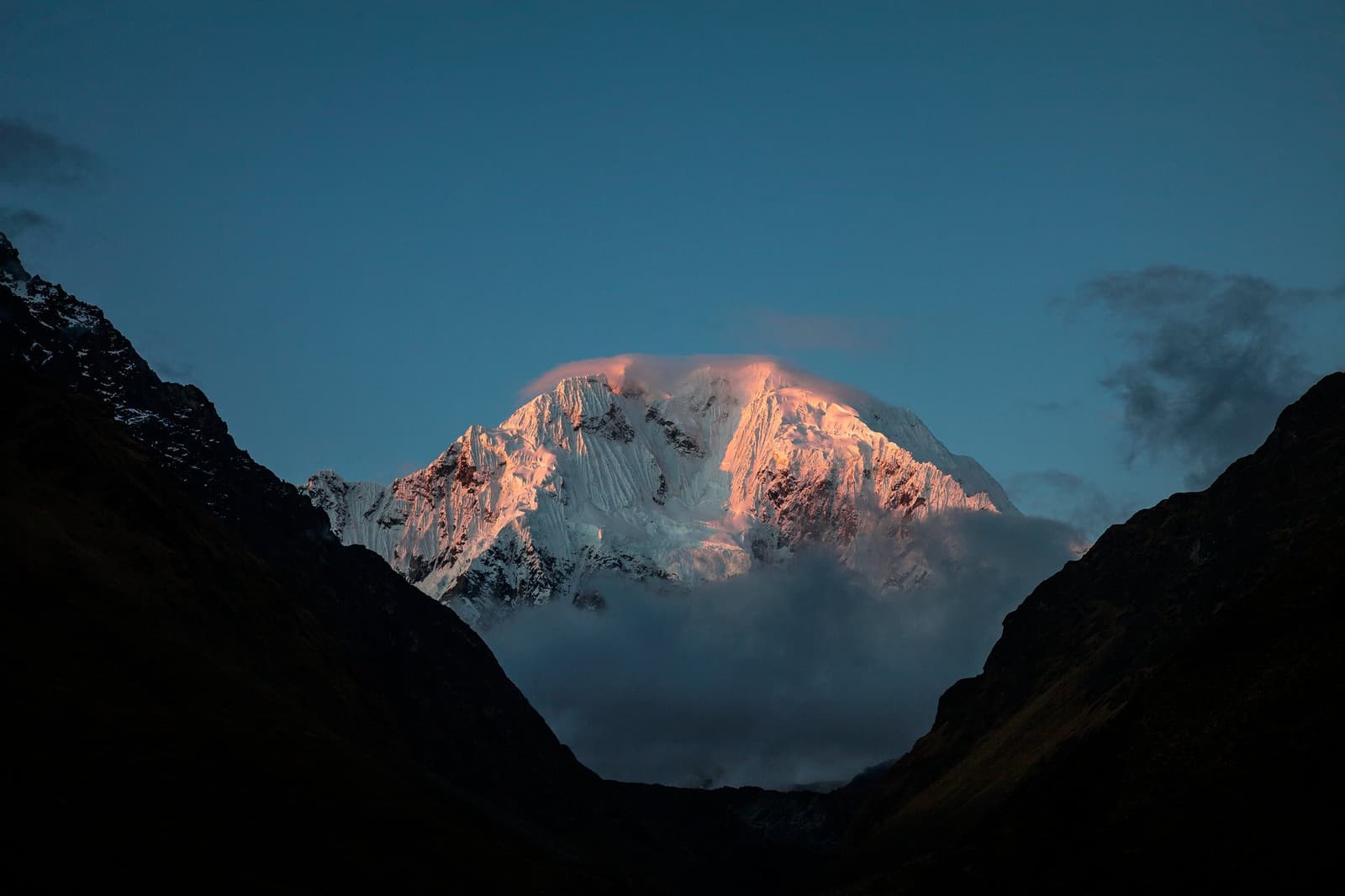 First light hits the ice before the rest of the world is even awake. A snow-capped Andean peak glowing pink and amber at sunrise on the luxury Salkantay trek, framed by dark valley ridges with low cloud clinging to its base