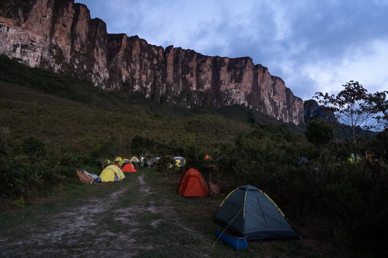 Acampamento pronto, a parede de pedra bem ali, e a subida é amanhã. Barracas coloridas armadas ao entardecer em uma clareira gramada durante uma expedição ao Monte Roraima, com a imponente parede de arenito avermelhada sob um céu nublado ao fundo