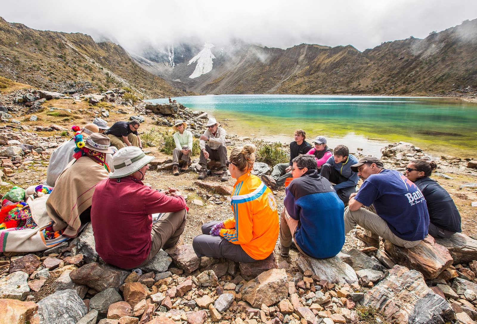 The Pachamama ceremony asks you to sit, listen, and mean it. A group of travelers and a local guide in traditional Andean poncho seated in a circle on the rocky shore of turquoise Humantay Lake during a salkantay tour, with snow-dusted peaks and low clouds rising behind the water