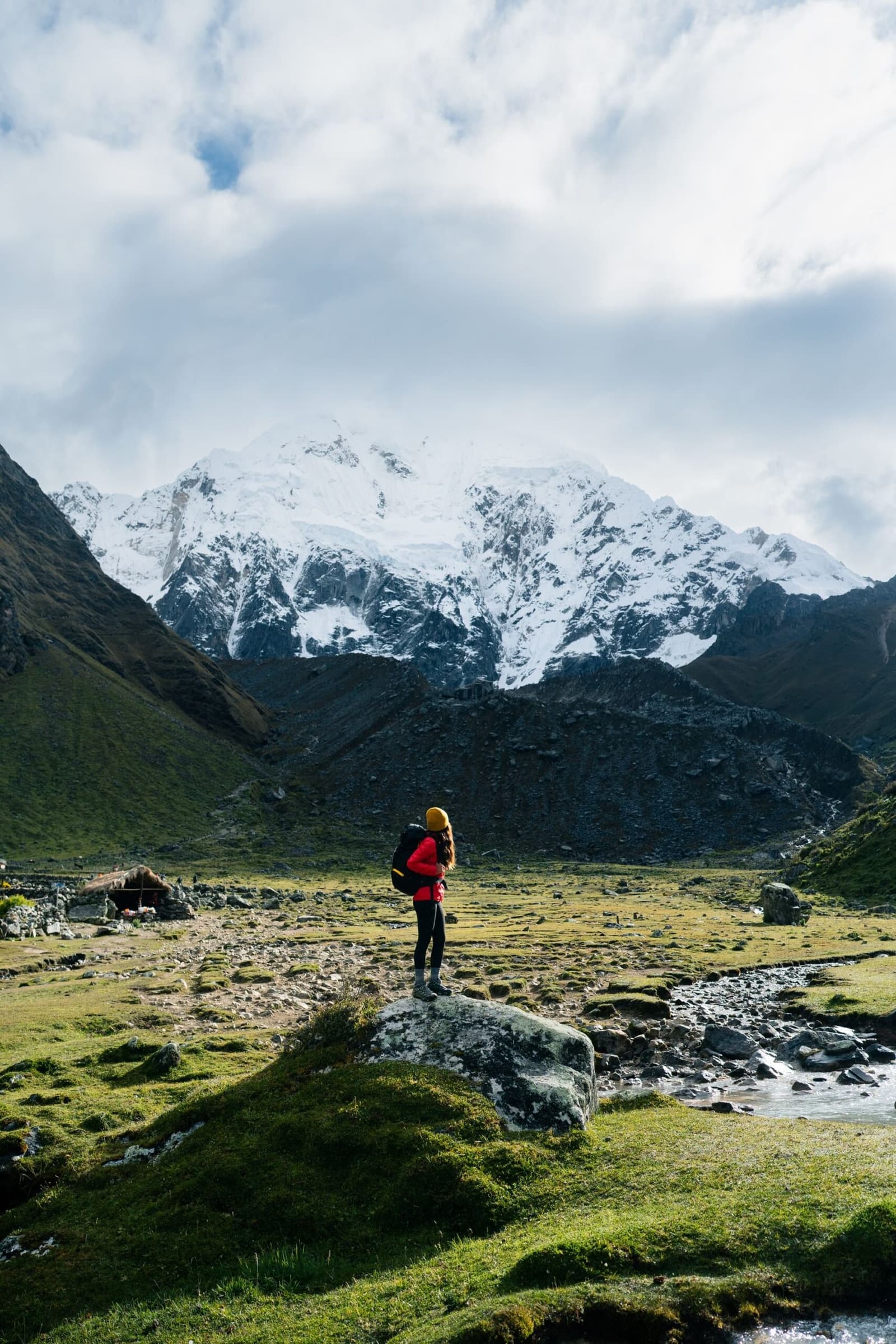 One rock, one enormous glacier, and a very good reason to pause. A solo hiker in a red jacket and yellow beanie stands on a boulder during an Andes mountains tour, facing a massive snow-capped peak above a wide high-altitude valley with a small stream and distant shelter