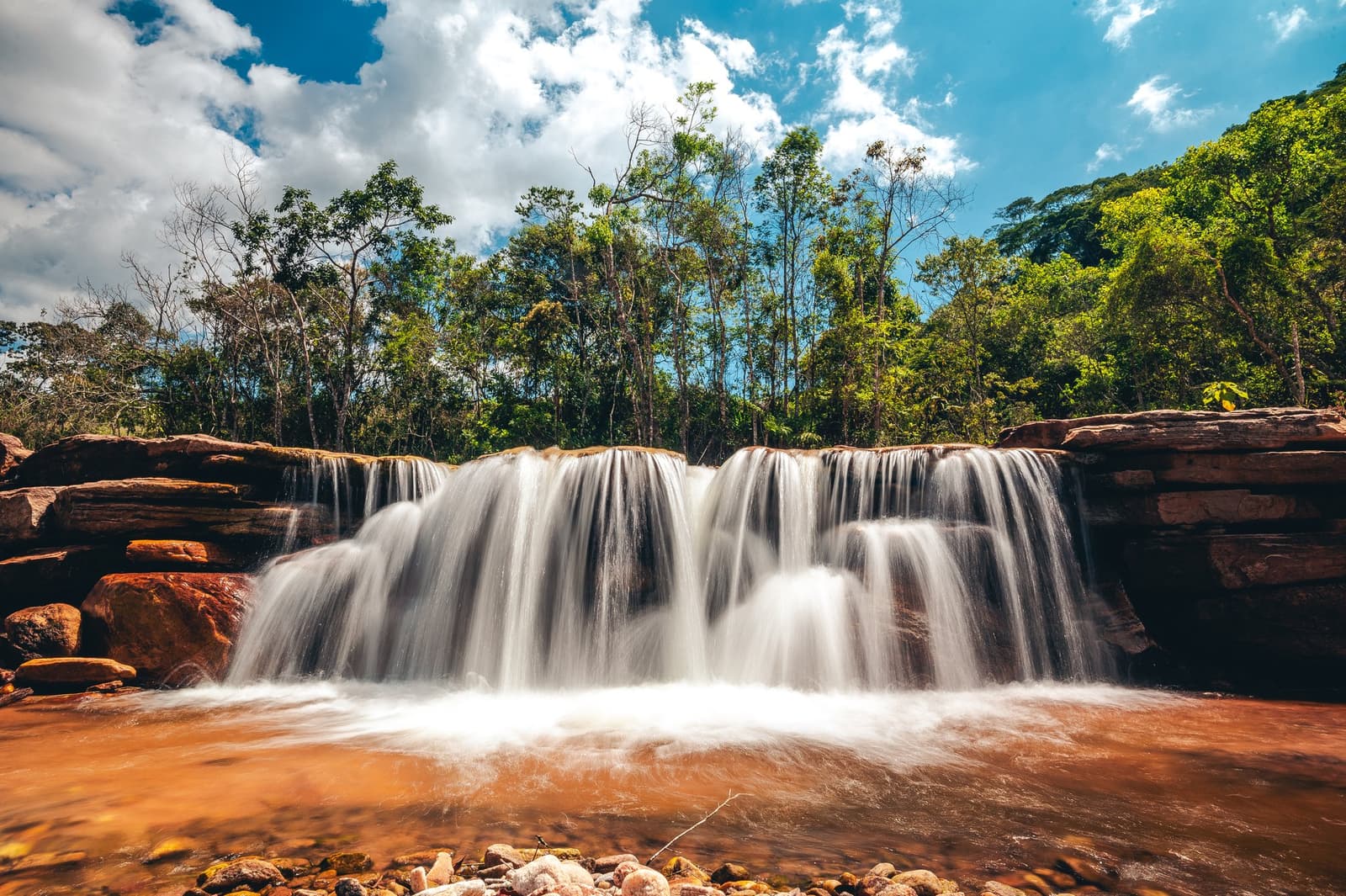Roter Sandstein, weißes Wasser, und kein Mensch weit und breit. Photo: Igor Tibirica Ein breiter Wasserfall, der über roten Sandsteinfelsen in ein kupferfarbenes Becken stürzt, umgeben von grünem atlantischen Regenwald unter einem teils bewölkten Himmel im Ibiti Project in Minas Gerais.