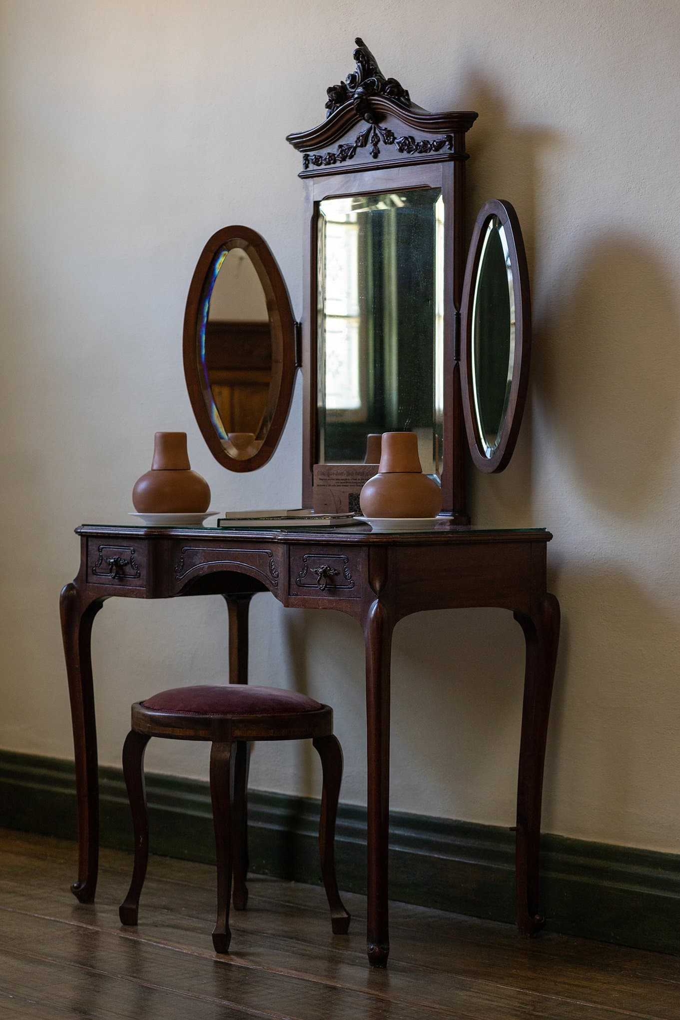 Un meuble qui a vu passer des générations, toujours debout, toujours élégant. Photo: Henrique Mangeon Coiffeuse en bois sombre ornée d'un miroir à fronton sculpté, deux miroirs ovaux latéraux, des vases en terre cuite et un tabouret en velours dans une chambre du lodge Ibiti Project