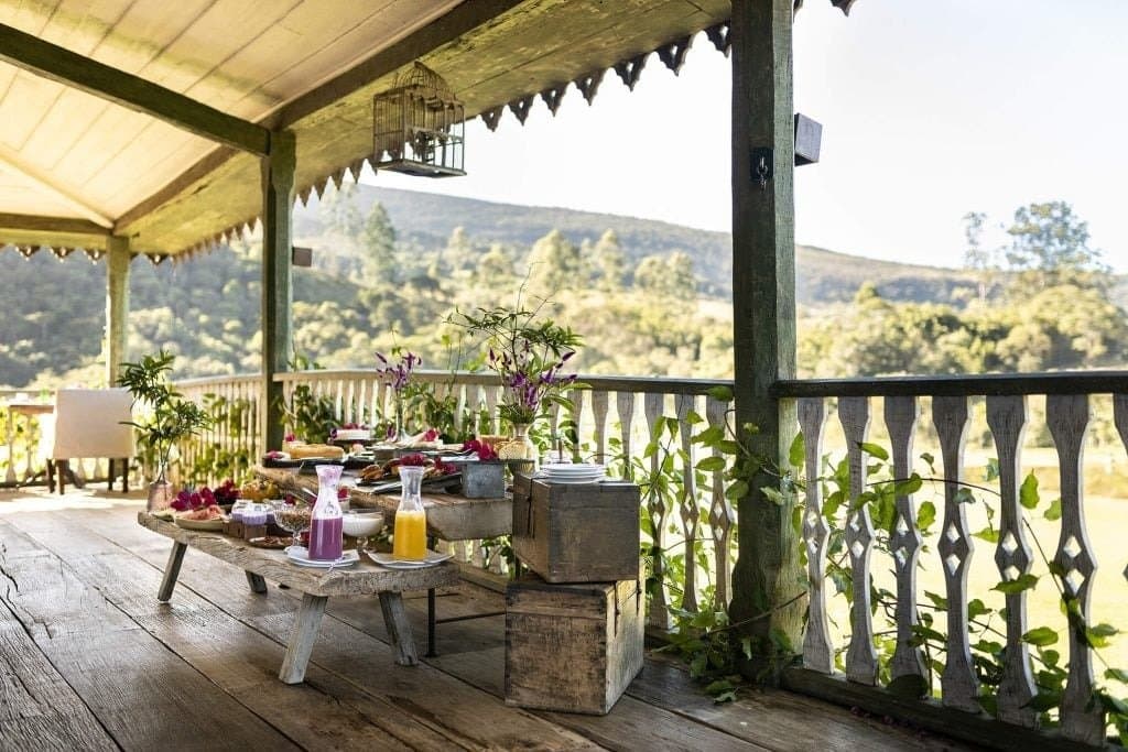 Frühstück mit Aussicht, die keinen weiteren Plan braucht. Eine rustikale Holzveranda der Engenho Lodge im Ibiti-Projekt mit einem Frühstücksbuffet aus frischen Säften, Früchten und Blumen, dahinter bewaldete Hügel