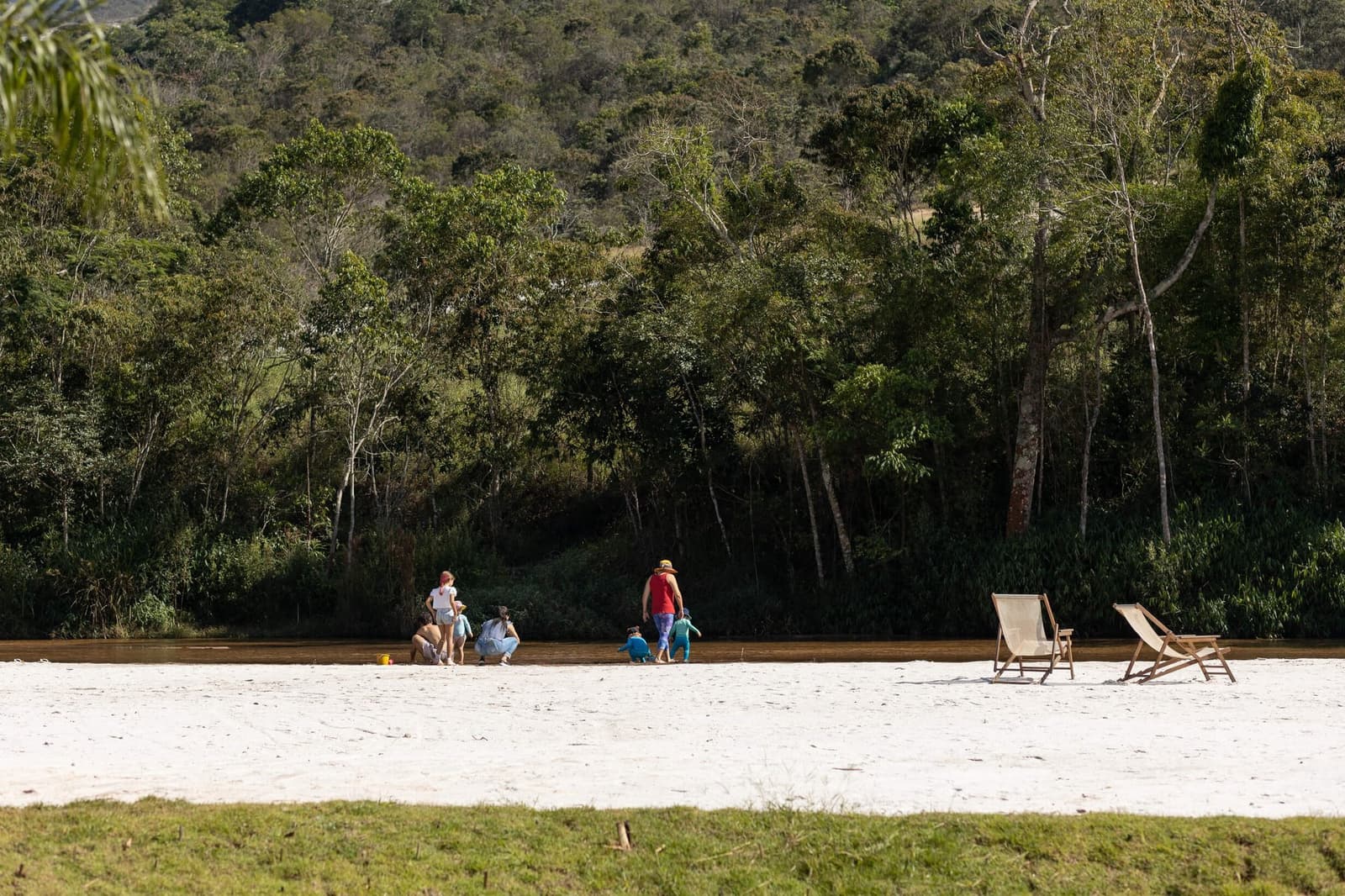 Du sable, une rivière tranquille, et des enfants qui n'ont plus rien à demander. Des familles et des enfants jouent au bord d'une plage de sable sur une rivière dans le cadre de l'Ibiti Project, avec des chaises longues en bois et une forêt atlantique dense sur la colline en arrière-plan