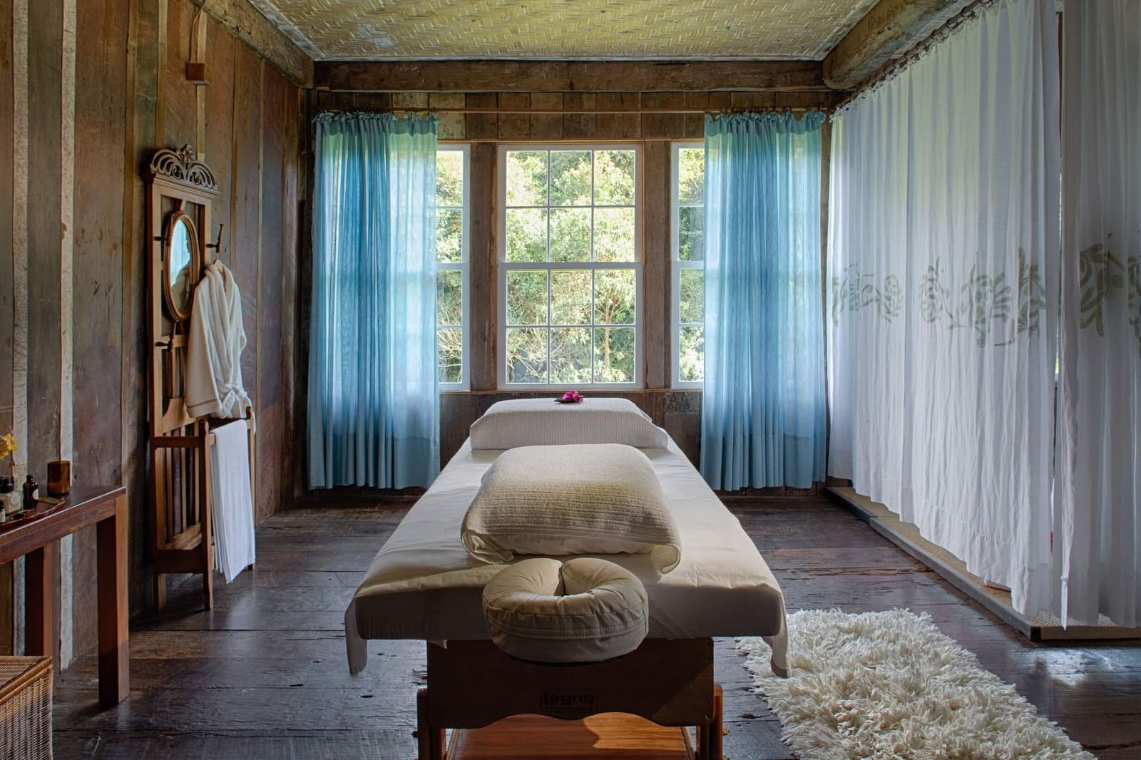 Le temps ralentit ici, le temps d'un soin, la forêt juste derrière la vitre. Une salle de soins apaisante au Spa Raízes de l'Ibiti Project, avec une table de massage rembourrée, des oreillers moelleux, des rideaux voilés bleu clair, des murs en bois rustique et un peignoir blanc accroché à un porte-manteau vintage près d'une fenêtre donnant sur la canopée