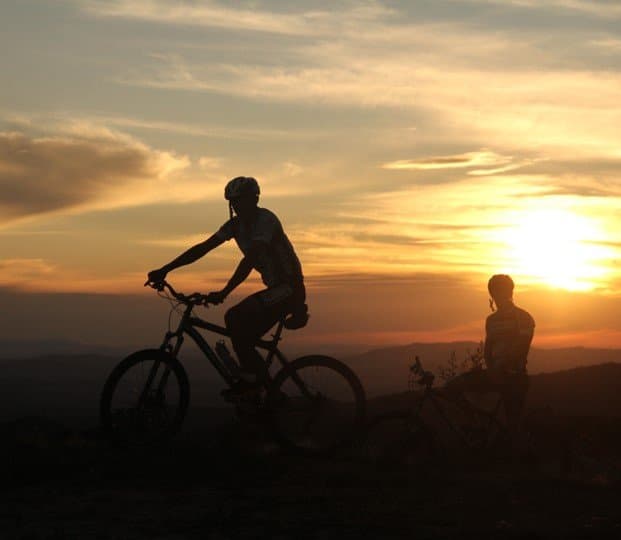 Zwei Fahrer, ein Kamm, das Ende eines Tages, der es verdient hat. Zwei Radfahrer als Silhouetten vor einem warmen orangefarbenen Sonnenuntergangshimmel auf einem Höhenkamm beim Ibiti Project