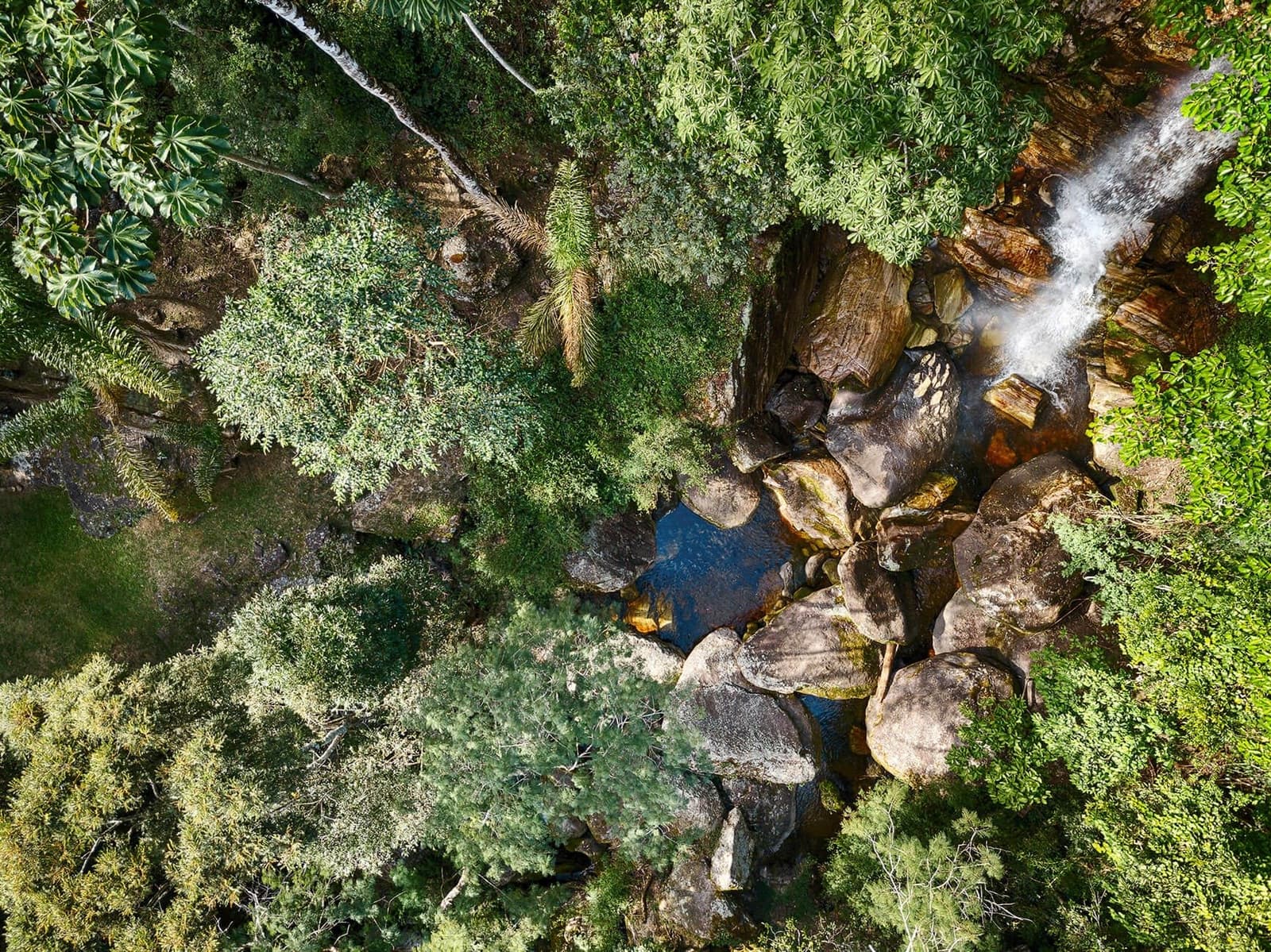 Von oben sieht man, wie das Wasser seinen eigenen Weg findet. Luftaufnahme eines Wasserfalls, der über große gerundete Felsen in ein dunkles Naturbecken stürzt, umgeben vom dichten Blätterdach des Atlantischen Waldes im Ibiti Project
