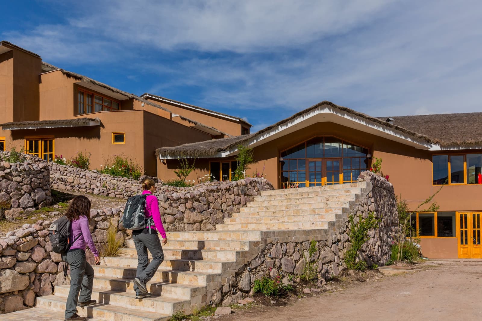 Packs on, altitude in the air, and a warm lodge waiting at the top of those steps. Two travelers with backpacks climbing stone steps toward a thatched-roof Andean lodge on a luxury Machu Picchu tour, with adobe walls and yellow-framed windows under a blue sky