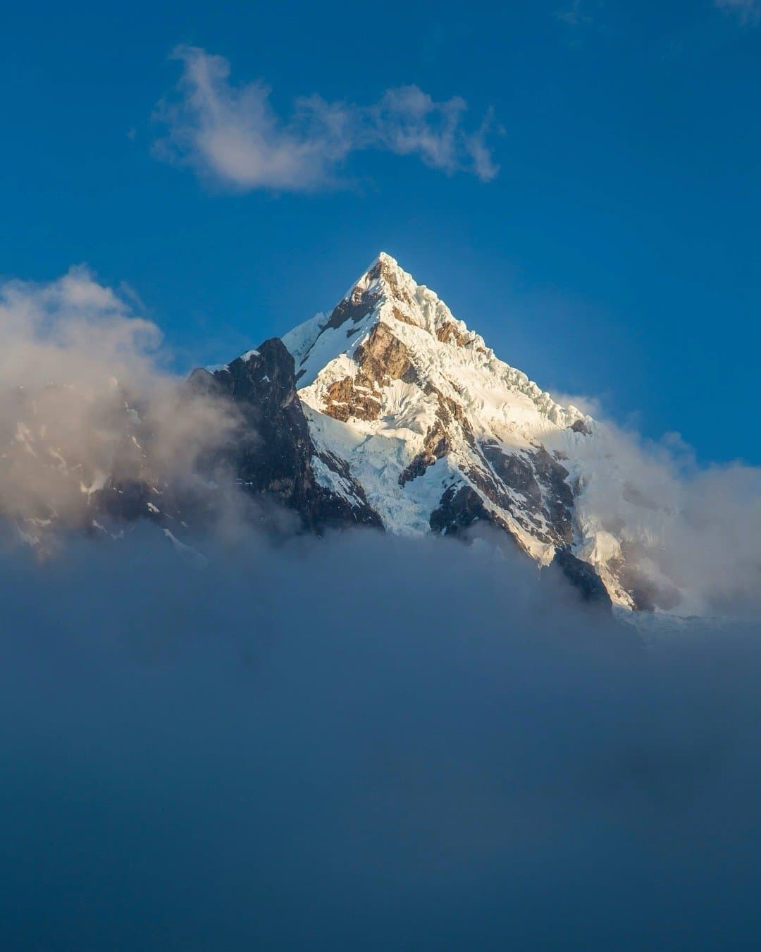 The mountain decides when it shows itself. Today, it was generous. A sharp snow-covered mountain peak emerging through a thick sea of clouds on a luxury Machu Picchu tour, lit by warm golden light against a deep blue sky