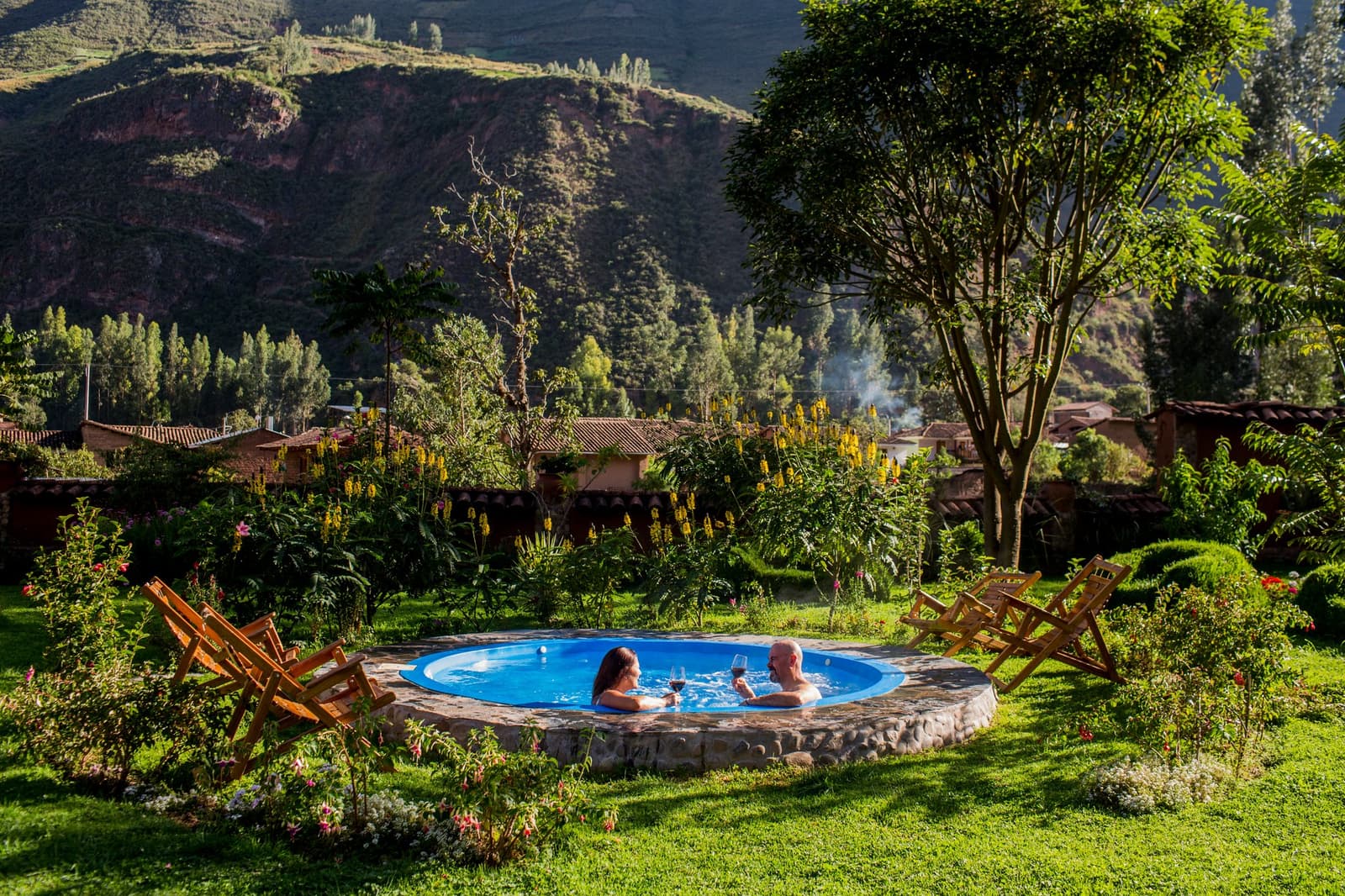 Wine, warm water, and the Andes right outside the garden fence. A couple toasting with red wine in a round stone-edged outdoor hot tub surrounded by a lush garden, with Andean hills in the background, on a luxury Machu Picchu tour