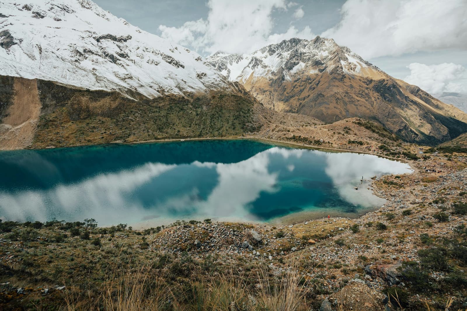 Cold water, cloud mirror, a tiny red jacket somewhere on the shore. A turquoise high-altitude glacial lake reflecting clouds, surrounded by arid rocky terrain and snow-capped Andean peaks on a luxury Machu Picchu tour