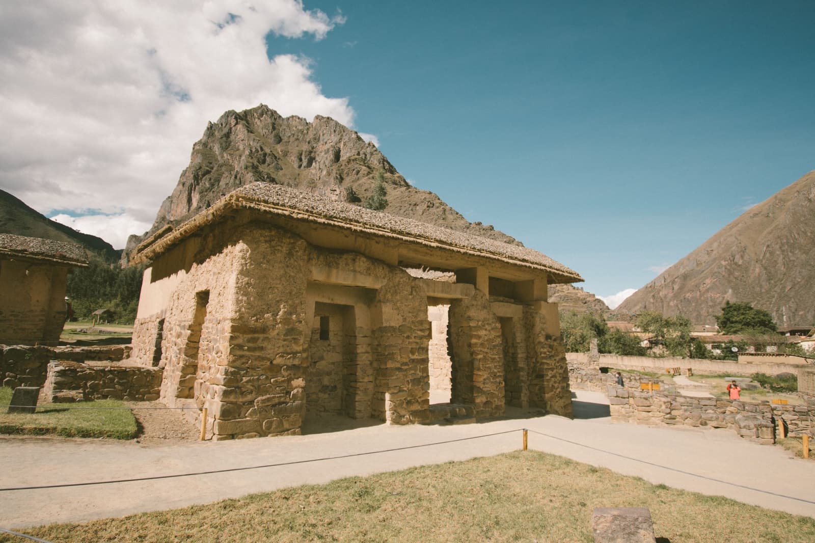 Stone fitted so well it has outlasted every government since. Restored Inca stone building with a thatched roof at Ollantaytambo archaeological site, part of a luxury Machu Picchu tour, with a jagged rocky peak rising directly behind it under a blue sky