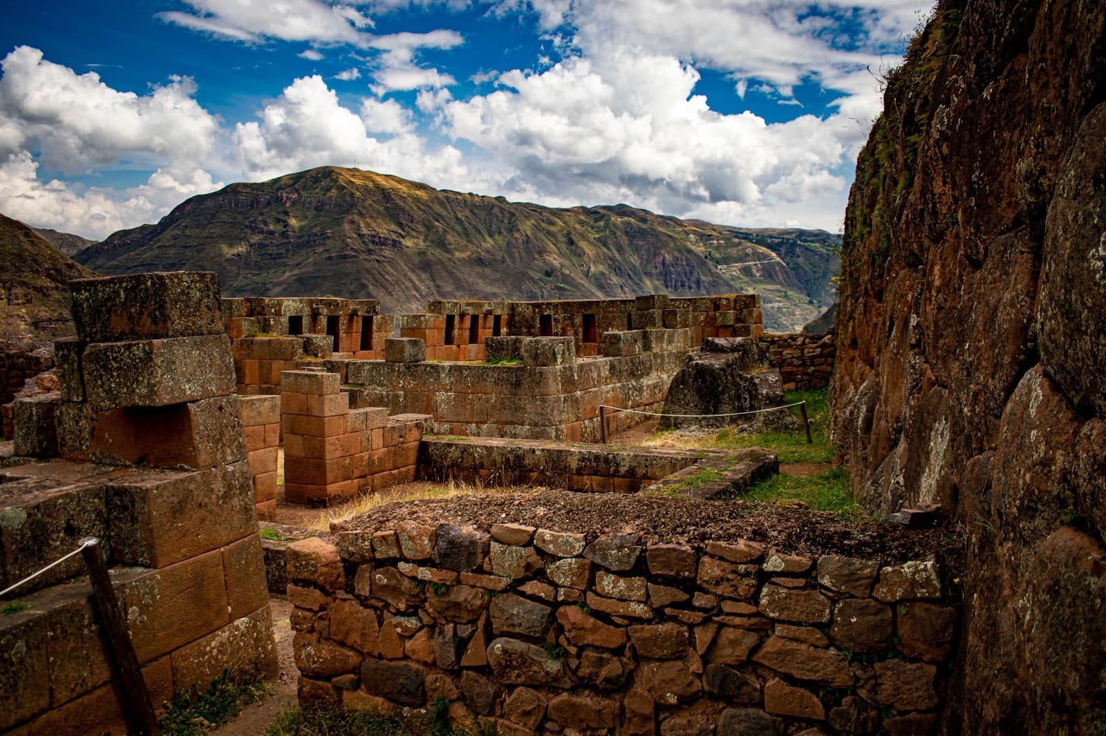 Stone cut this precisely, and they did it without a blueprint in sight. Finely fitted red stone walls and multi-level platforms of Inca ruins on a luxury Machu Picchu tour, with Andean mountain ridges and blue sky in the background