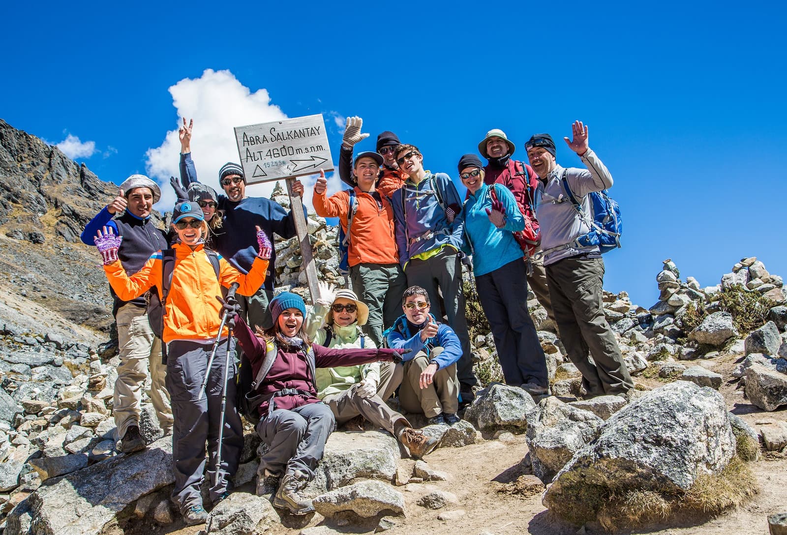 4,600 metres up, everyone's grinning. Turns out altitude agrees with good company. A group of excited trekkers celebrating at the Abra Salkantay sign at 4,600 m on the luxury Salkantay trek, arms raised and grinning under a deep blue Andean sky with rocky peaks behind them