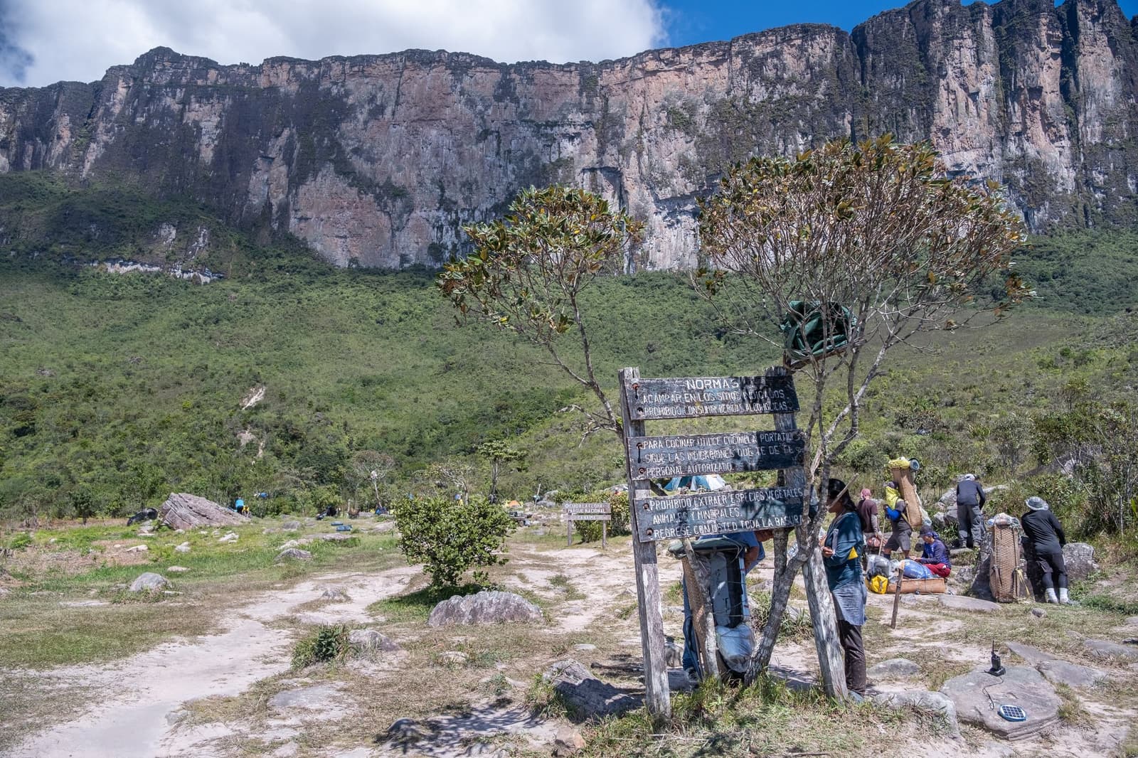 Normas na placa, cestos no ombro, e a parede já não deixa dúvida nenhuma. Uma placa de madeira com 'Normas' está fixada no acampamento base de uma expedição ao Monte Roraima, enquanto carregadores preparam cestos tradicionais e a parede vertical do tepui se ergue ao fundo
