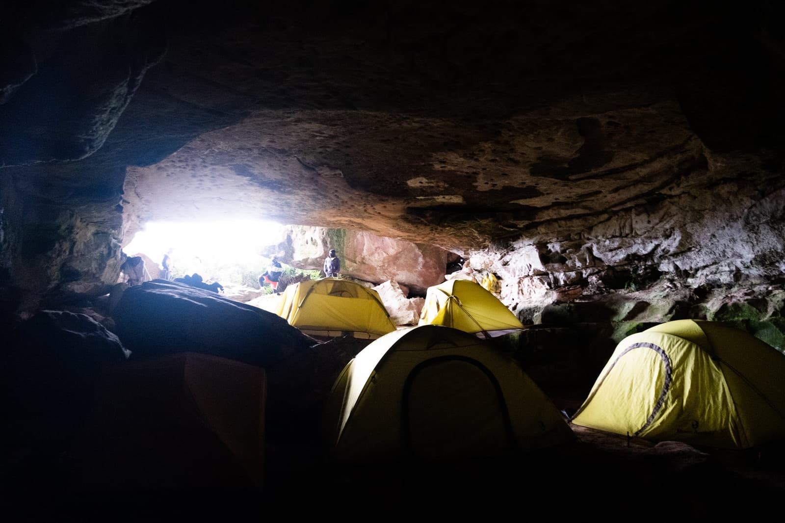 Teto de pedra, sem estacas necessárias. O Coati Camp cumpre o que promete. Quatro barracas cúpula amarelas armadas dentro de um grande abrigo sob rocha durante uma expedição ao Monte Roraima, com luz do dia e silhuetas de caminhantes visíveis pela abertura baixa da caverna
