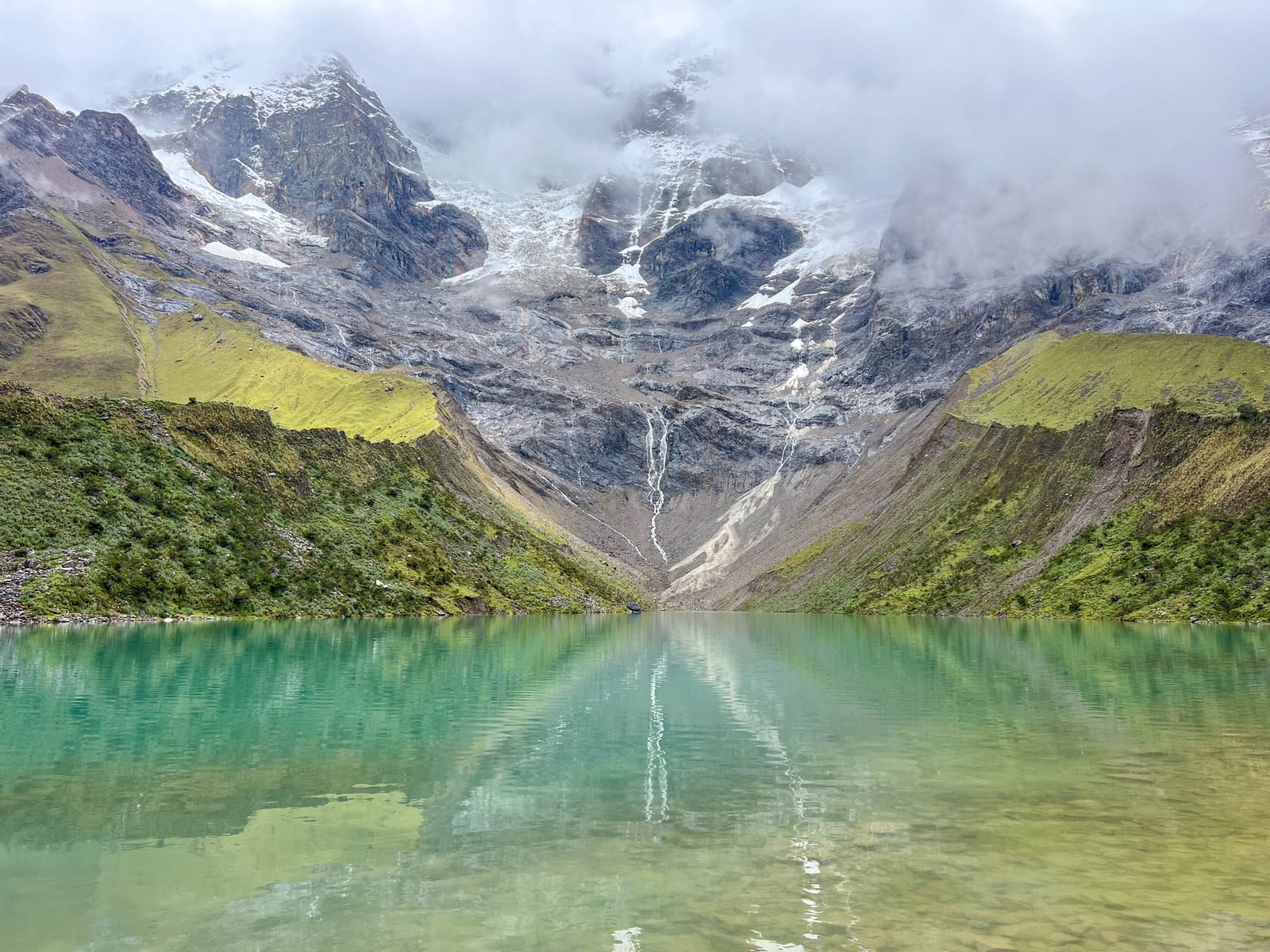Glacier melt, mineral water, that color. Worth every step of the climb. Photo: Mystockimages Turquoise glacial waters of Humantay Lake reflecting a snow-streaked rocky mountain face flanked by green Andean hillsides on a salkantay tour, with mist drifting across the peaks above