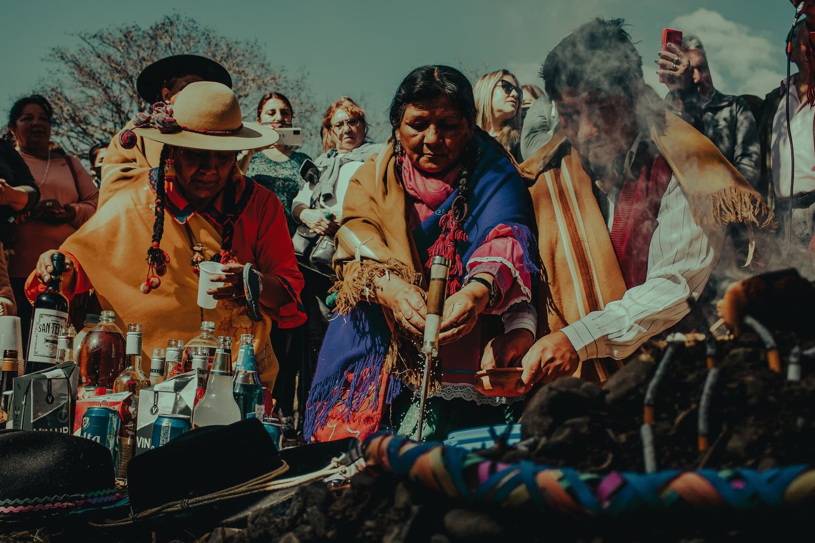 Smoke, libations, and a quiet word with Pachamama before the road home. Indigenous women in colorful ponchos and traditional hats conduct a Pachamama ceremony on this salkantay tour, pouring libations over a ritual offering table with bottles, candles, and sacred objects while smoke rises and a crowd of onlookers gathers around them