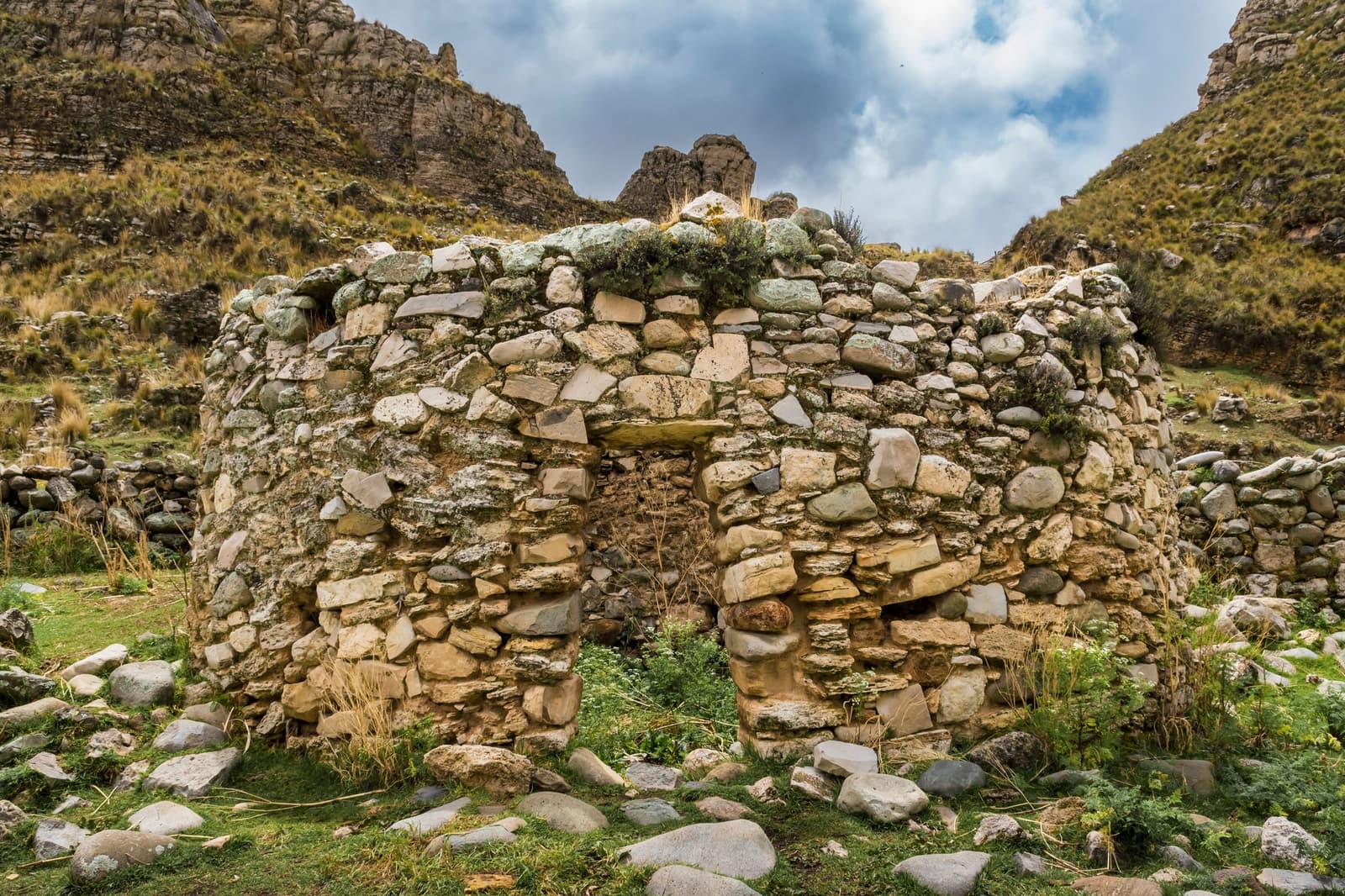 Centuries of Andean weather, and these walls still have something to say. Photo: Gilmer Diaz Estela Roofless circular dry-stone wall ruins at the Quillarumiyoc archaeological site, with lichen-covered fieldstone construction, wild grasses growing at the base, and rugged Andean peaks rising under a cloudy sky on the salkantay tour