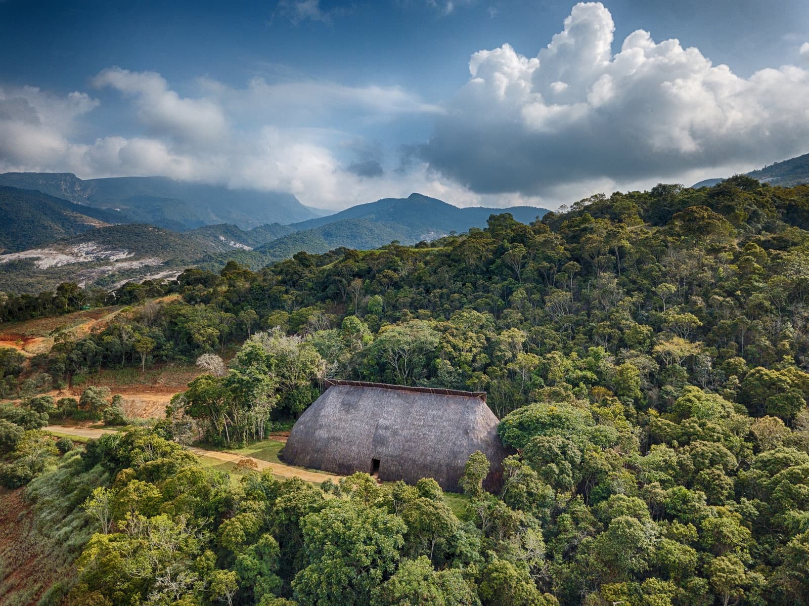 Sechstausend Hektar Wald, und ein Strohdach, das seinen Platz behauptet. Luftaufnahme einer großen traditionellen Oca-Struktur mit Strohdach, eingebettet in dichten Atlantischen Regenwald, mit bewaldeten Bergen und dramatischen Wolken im Hintergrund beim Ibiti Project in Minas Gerais