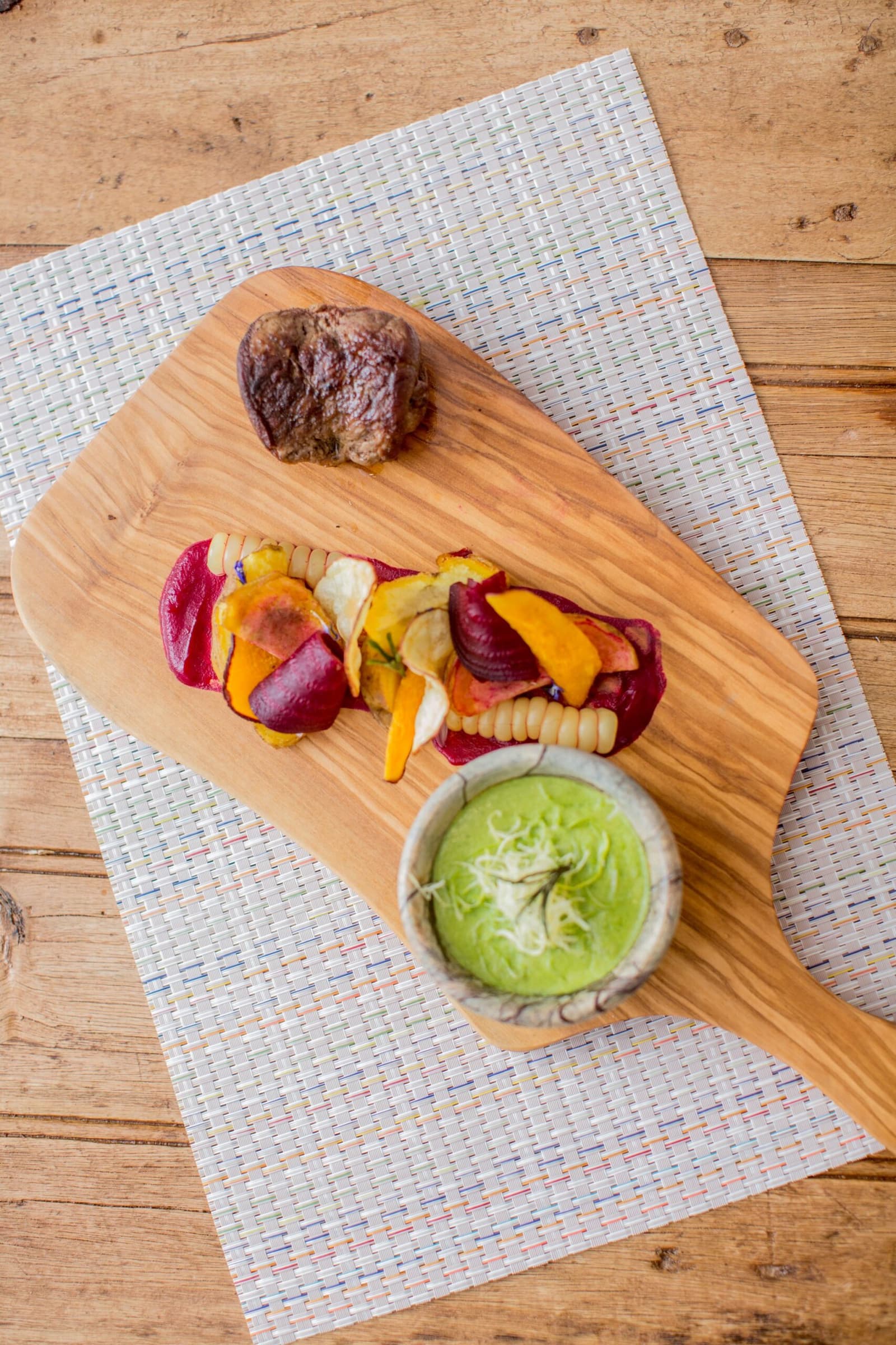 Rustic board, serious cooking. The Andes have their own idea of fine dining. A seared meat medallion served on a wooden board alongside colorful roasted vegetable chips and a small bowl of green herb sauce, part of a luxury Machu Picchu tour dining experience