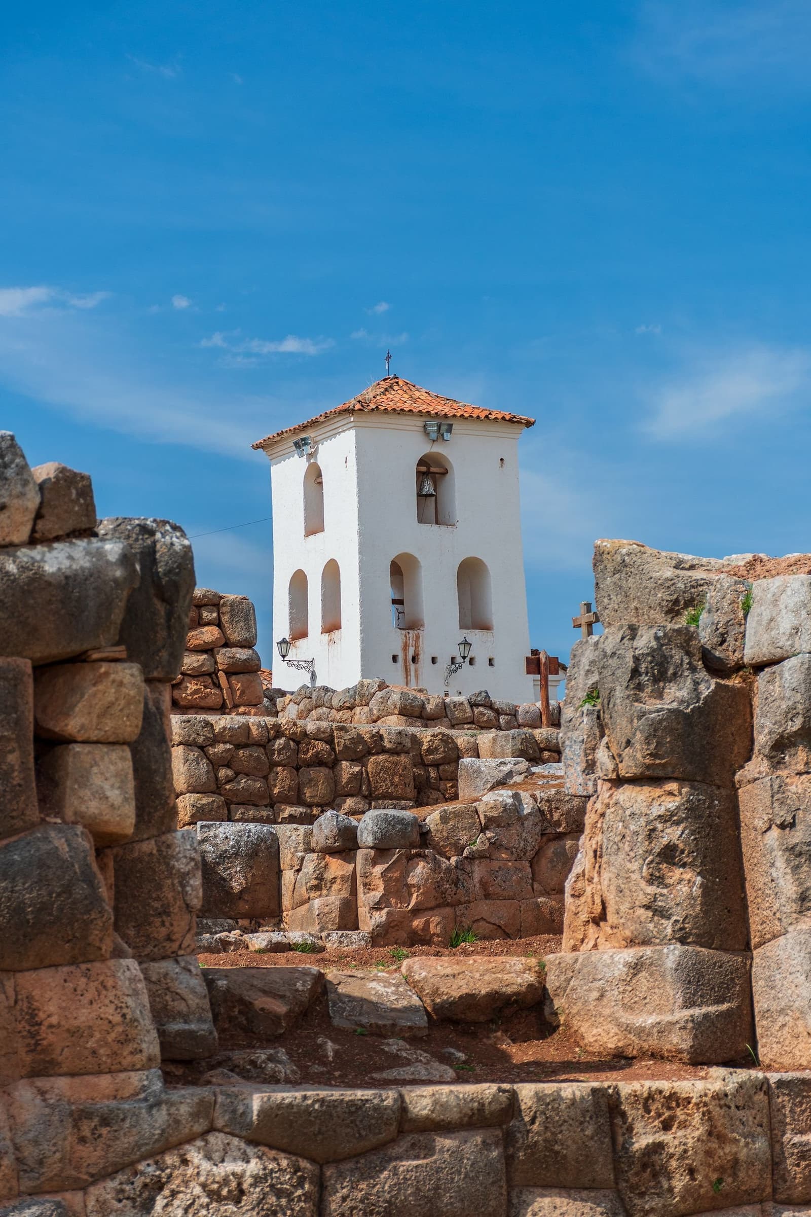 Two civilizations sharing the same hill, neither one asking permission. A white colonial bell tower with an orange terracotta roof rises above ancient Inca stone foundations and walls on this luxury Machu Picchu tour, framed against a clear blue sky with wooden crosses visible to the right