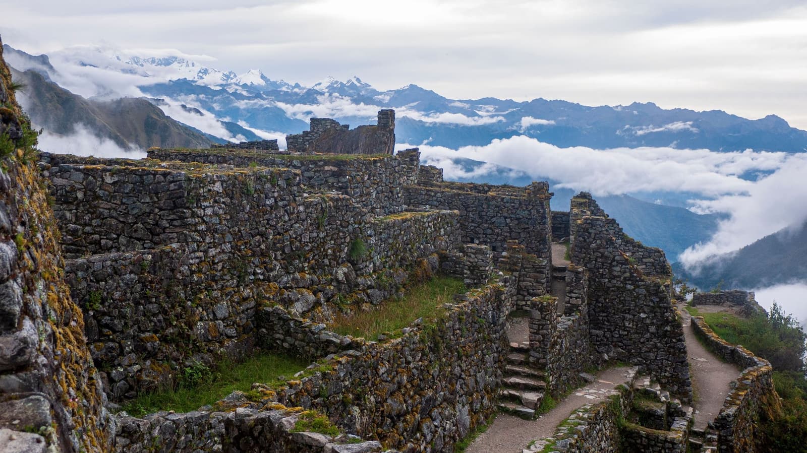 Clouds fill the valleys below, and the walls have been here long enough not to care. Moss-covered Inca stone ruins on a high ridge during a luxury Machu Picchu tour, with a stone stairway path winding through roofless walls and a sea of clouds rolling over snow-capped Andean peaks in the distance
