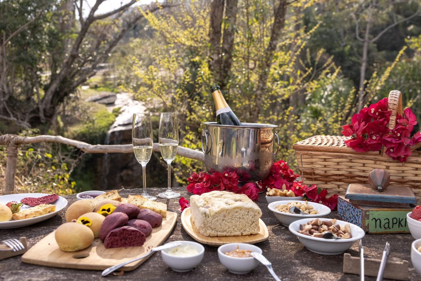 Frisch gebackenes Brot, eiskalter Schaumwein, Wald ringsum. So soll Mittagspause sein. Ein Picknicktisch beim Ibiti Project mit handwerklichem Brot, gemischten Nüssen, Dipsaucen, zwei Champagnergläsern und einer Flasche im Eiskühler, dekoriert mit roten Bougainvillea-Blüten und einem Weidenkorb, im Hintergrund Bäume und ein kleiner Wasserfall