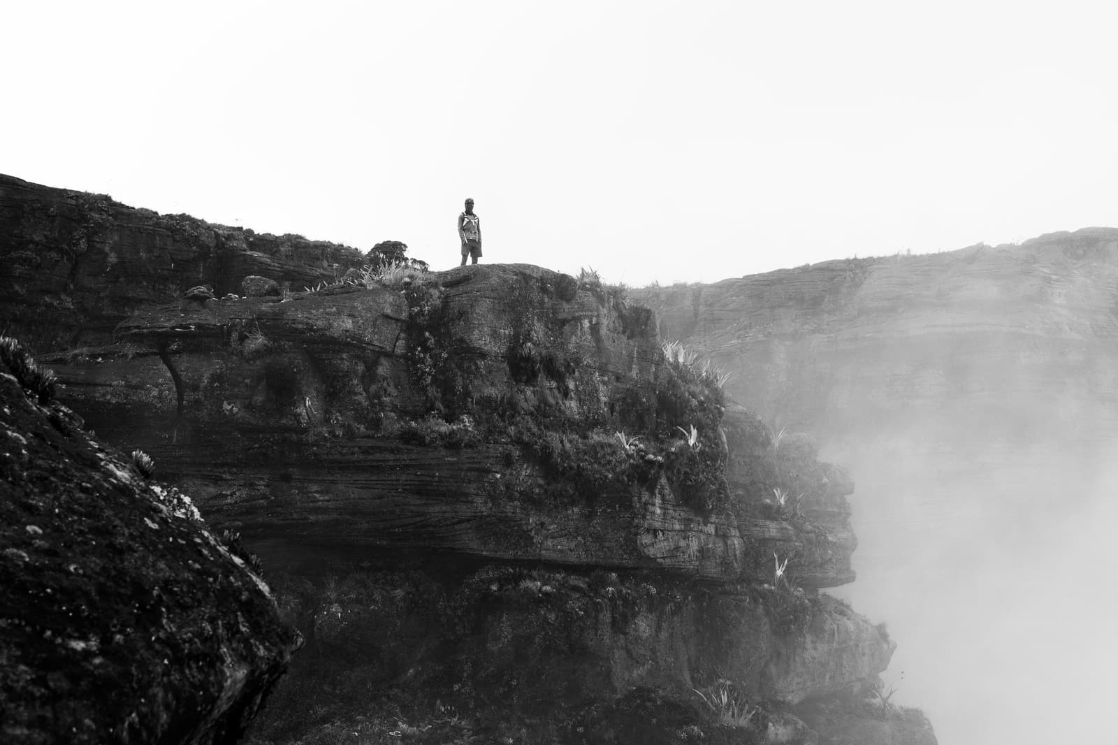 Você, o penhasco, e uma nuvem sem destino definido. Um caminhante solitário parado na borda de um penhasco de arenito coberto de musgo durante uma expedição ao Monte Roraima, com névoa densa subindo do cânion e paredes de rocha em camadas desaparecendo no branco
