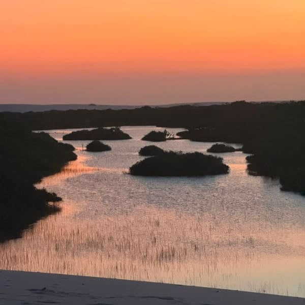 Orangefarbener Himmel spiegelt sich in einer Lagune mit Vegetationsinseln, von einer Düne in den Lençóis Maranhenses aus gesehen