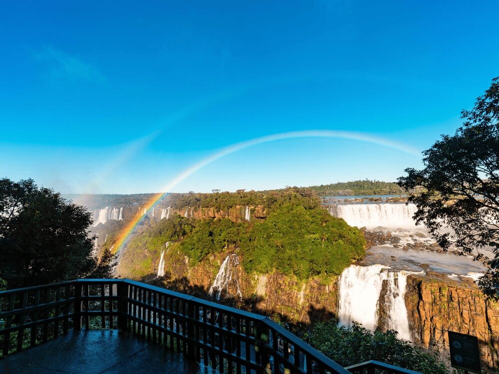 Photo: paulobaqueta Rainbow over Iguazu Falls seen from a viewpoint railing on a clear day