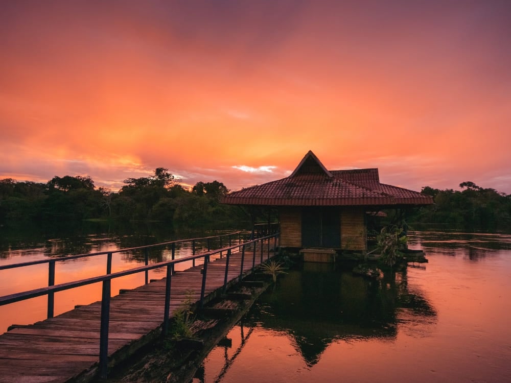 Photo: Gui Gomes Wooden walkway over the river leading to a stilted cabin under an orange sunset in the Amazon