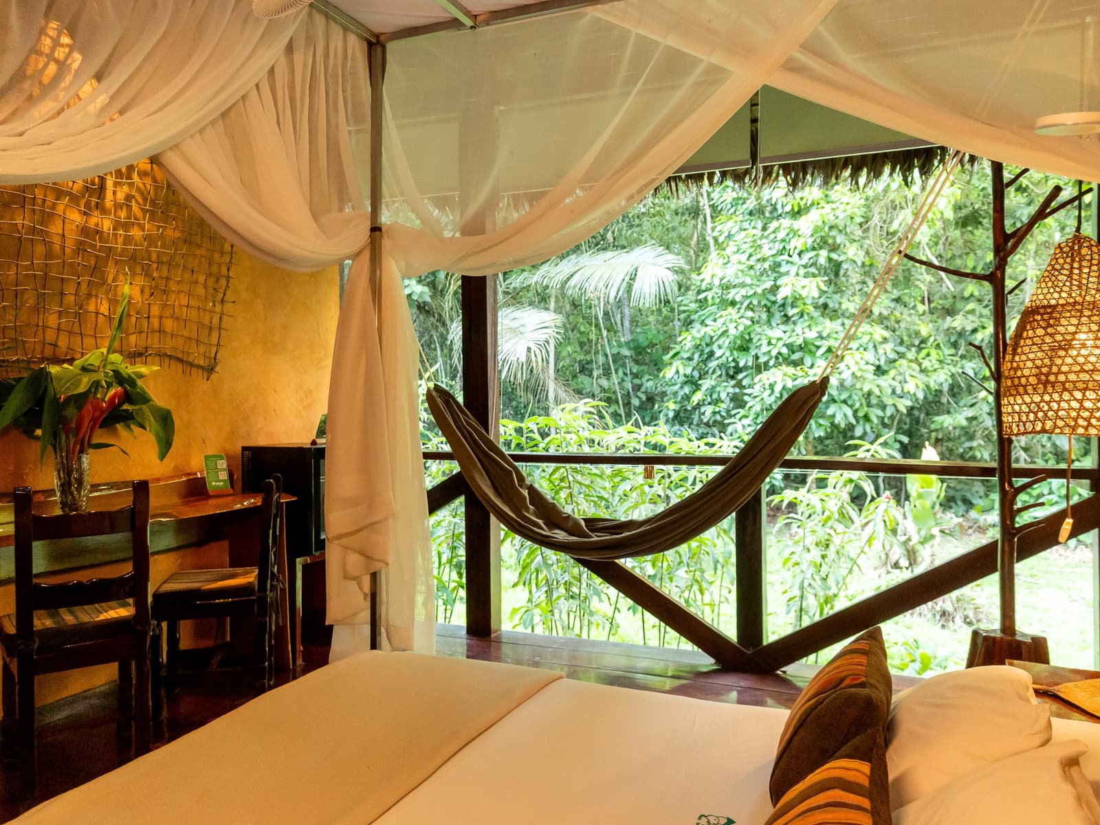 Bed, hammock, jungle, in that order, depending on the hour. Canopied bed with white mosquito netting inside a Posada Amazonas lodge room, with a hammock hanging on the open balcony overlooking dense rainforest green on this amazon tour from puerto maldonado