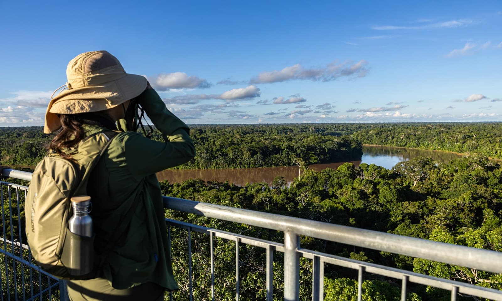 Binoculars up, the forest stretches further than you expect. A traveler with binoculars and a backpack looks out from the 40-metre canopy tower over an unbroken expanse of rainforest canopy and a winding brown river below on this amazon tour from puerto maldonado