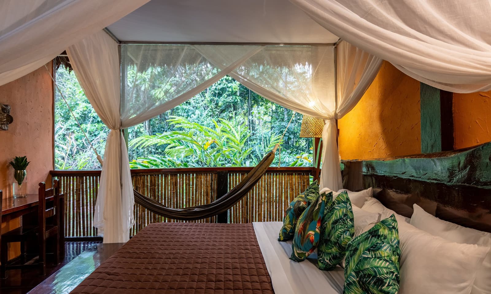 Your alarm clock here is a howler monkey, not a phone. Four-poster bed with white mosquito netting at Posada Amazonas, opening onto a bamboo balcony with a hammock and dense rainforest canopy beyond