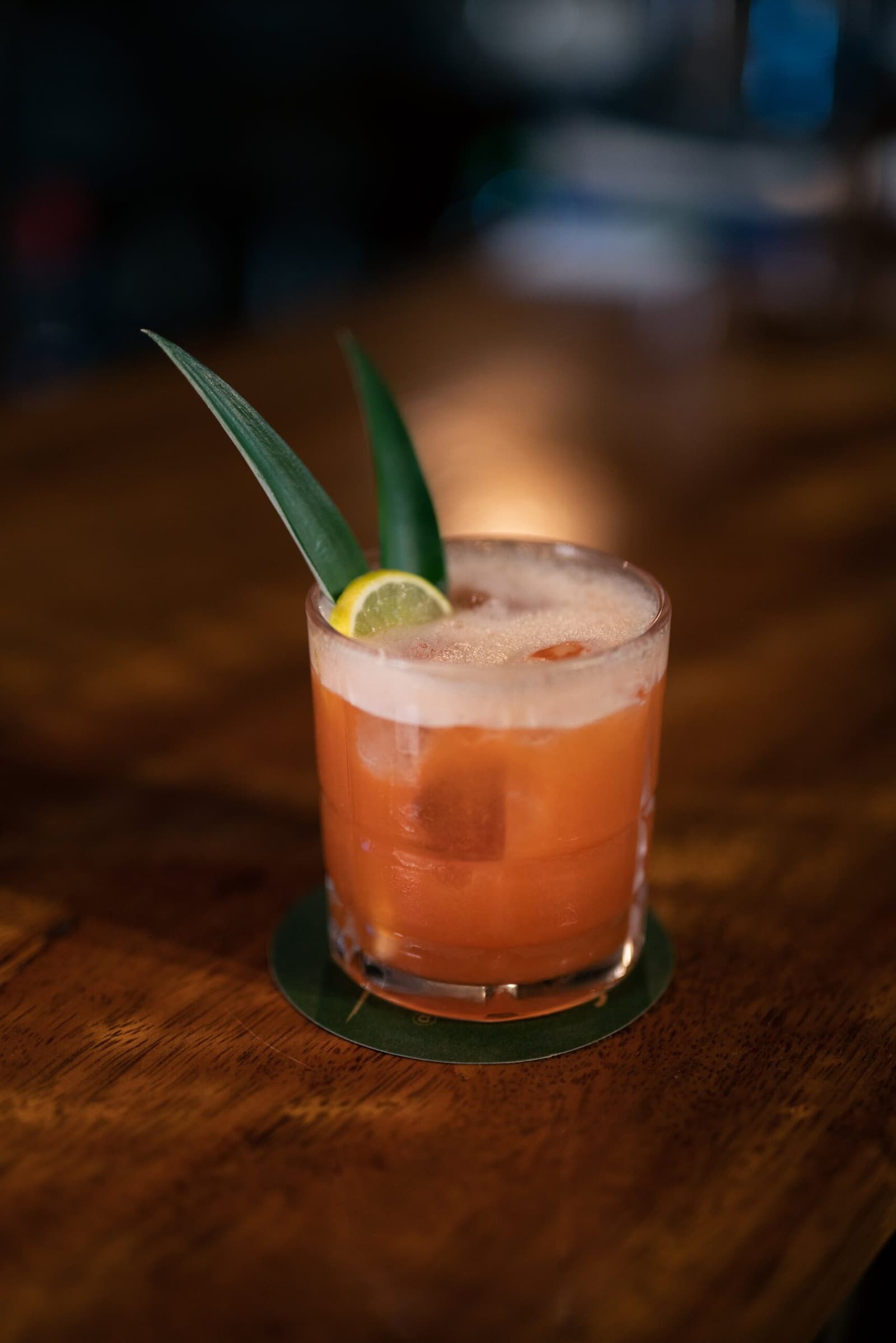 Jungle day done. This one earns its garnish. A frosted rocks glass filled with an amber-orange cocktail, garnished with a lime wedge and two long tropical leaves, resting on a green coaster on a warm wooden bar counter at Posada Amazonas lodge