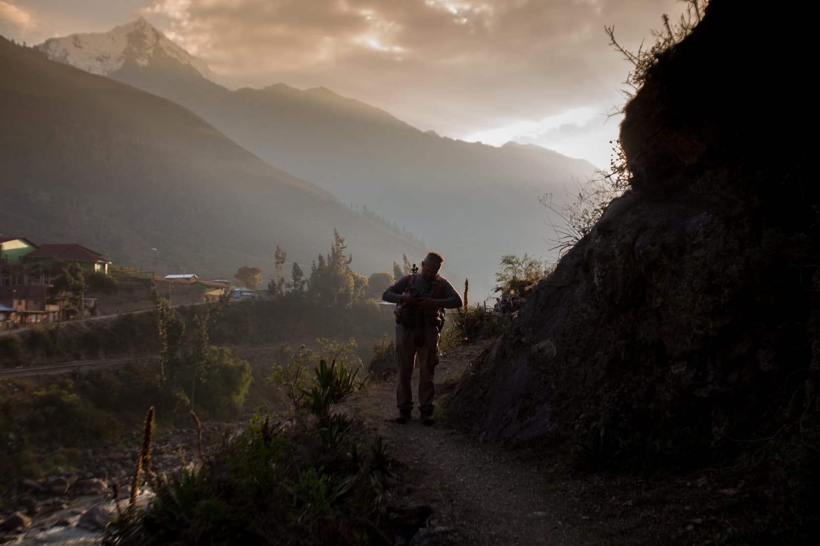 Primeira noite na trilha, e a montanha já tem algo a dizer. Um trekker solitário faz uma pausa em uma trilha estreita ao entardecer no trek Ancascocha, em silhueta contra um vale andino enevoado com picos nevados e uma pequena aldeia visível abaixo